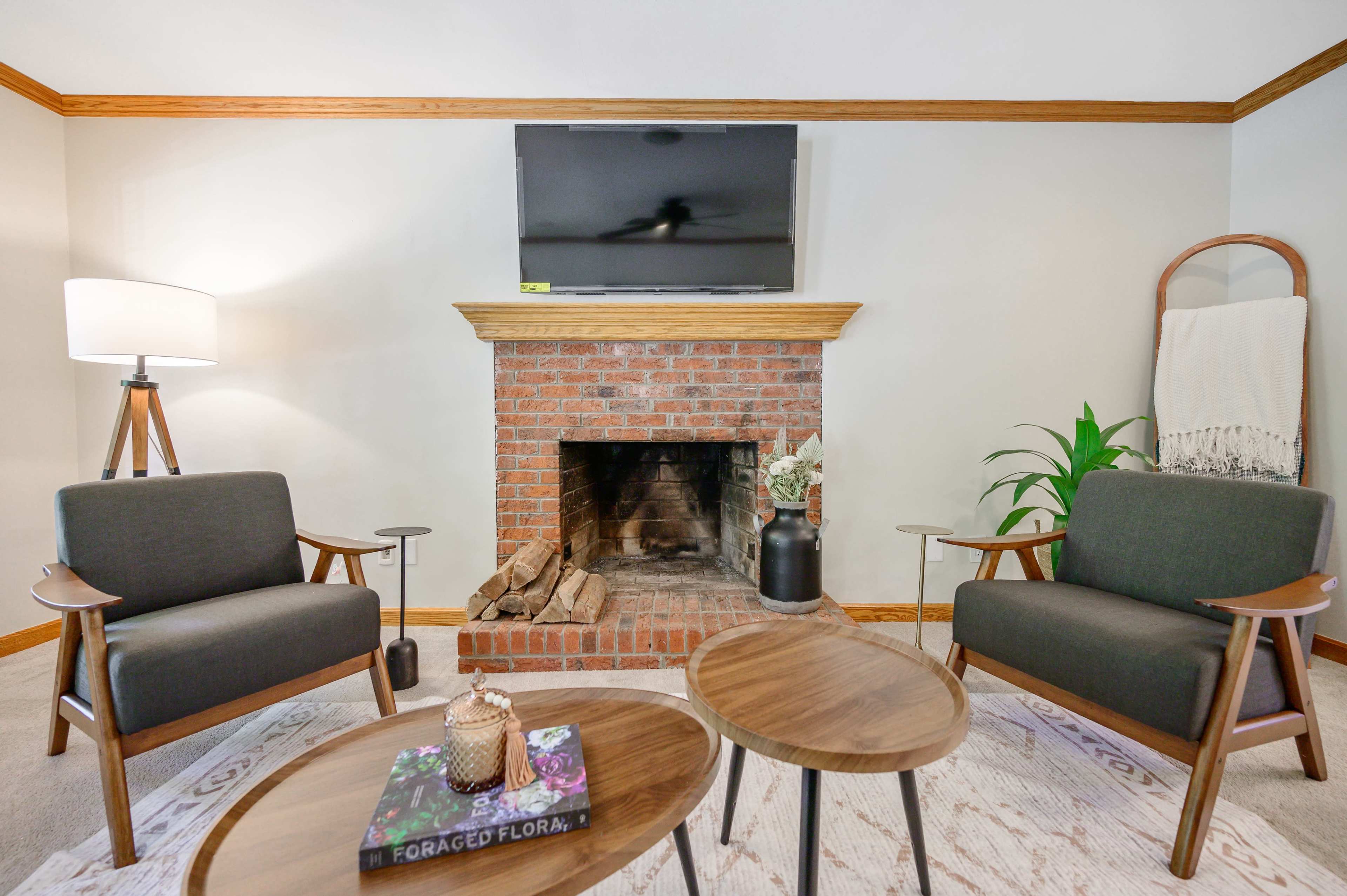 A cozy living room features two gray armchairs facing a brick fireplace with a television mounted above it, accompanied by two wood tables and a floor lamp.