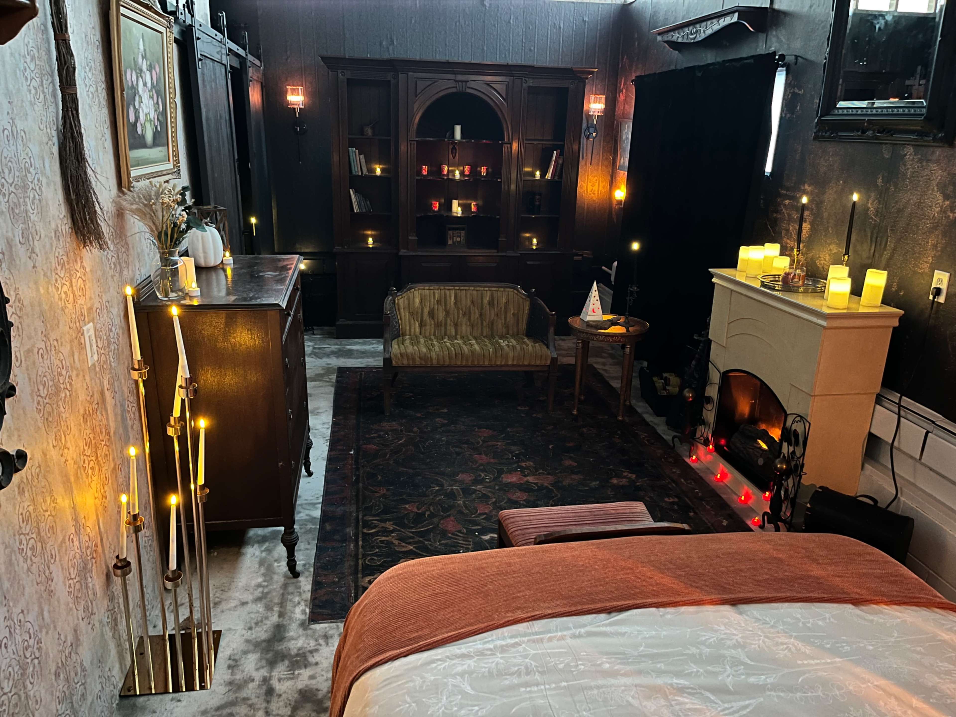 The image shows a dimly lit bedroom with a cozy sitting area, a vintage-style cabinet, and a decorative electric fireplace surrounded by candles.