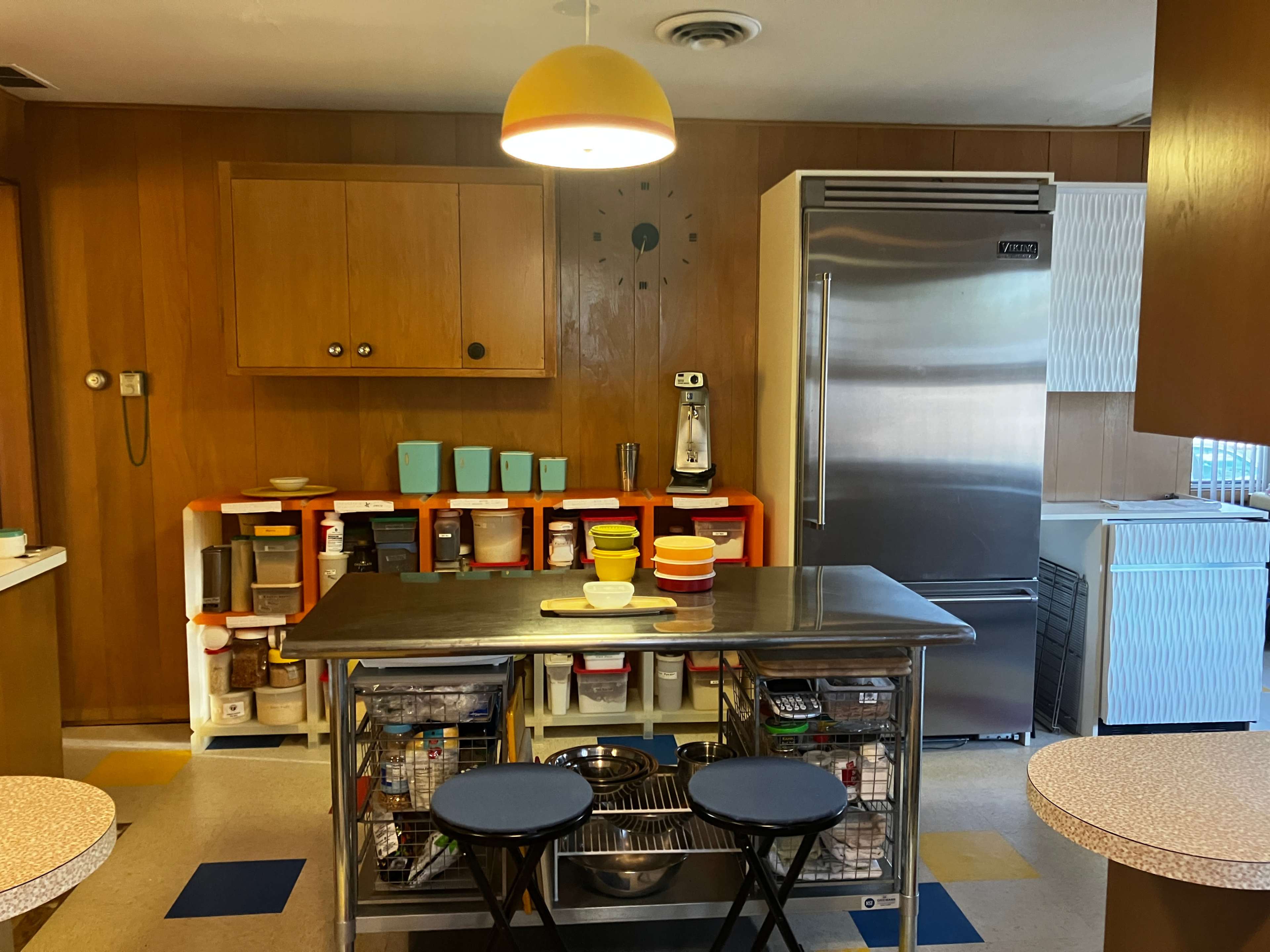 The image shows a kitchen with wooden paneling, a central kitchen island, and organized shelves containing various kitchen items.