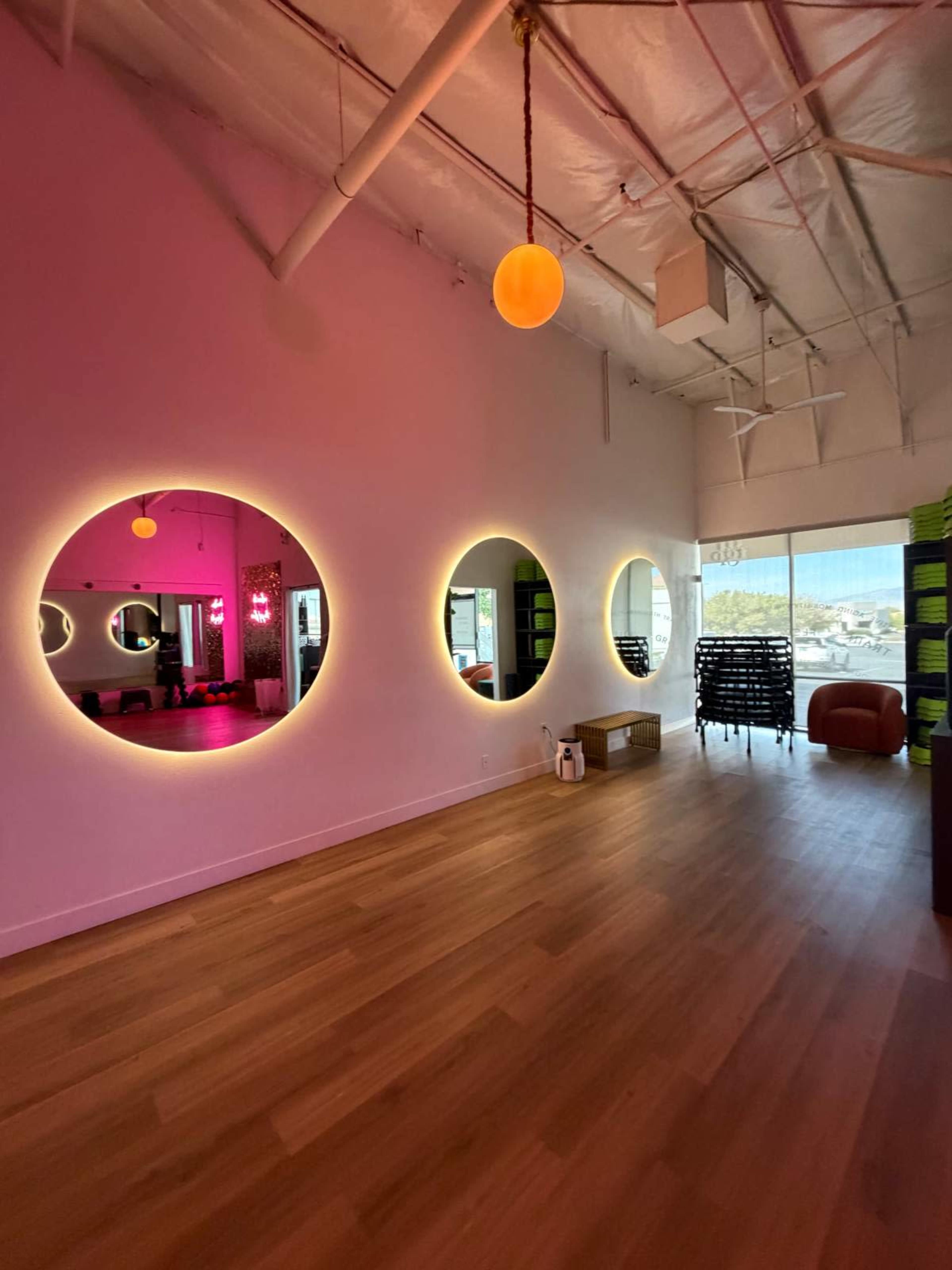 The image shows a spacious, well-lit room with four circular mirrors illuminated by soft light, featuring a wooden floor and a large window with a view outside.