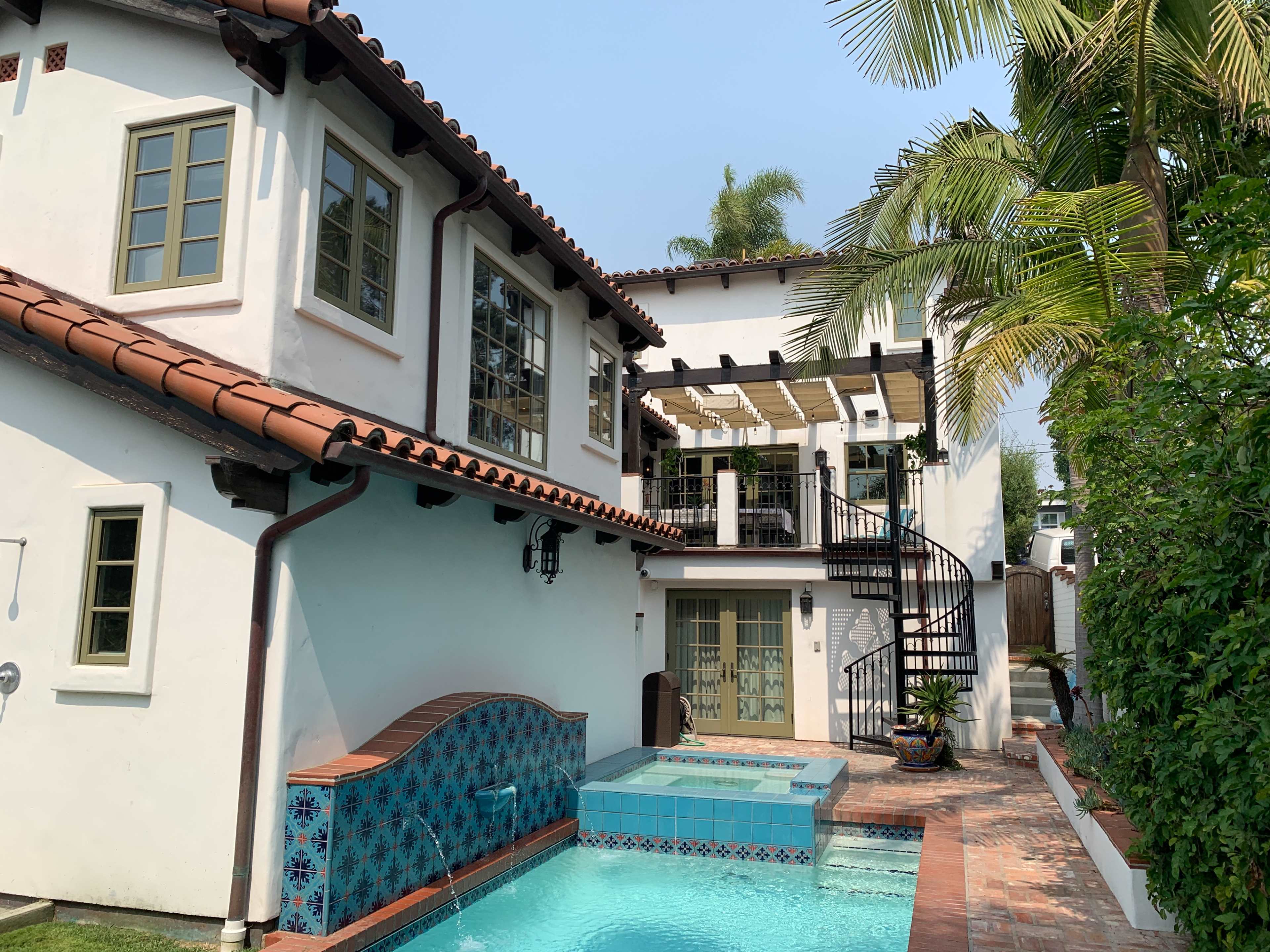 A two-story house with a tiled roof, a small swimming pool, and a spiral staircase leading to a balcony surrounded by palm trees.