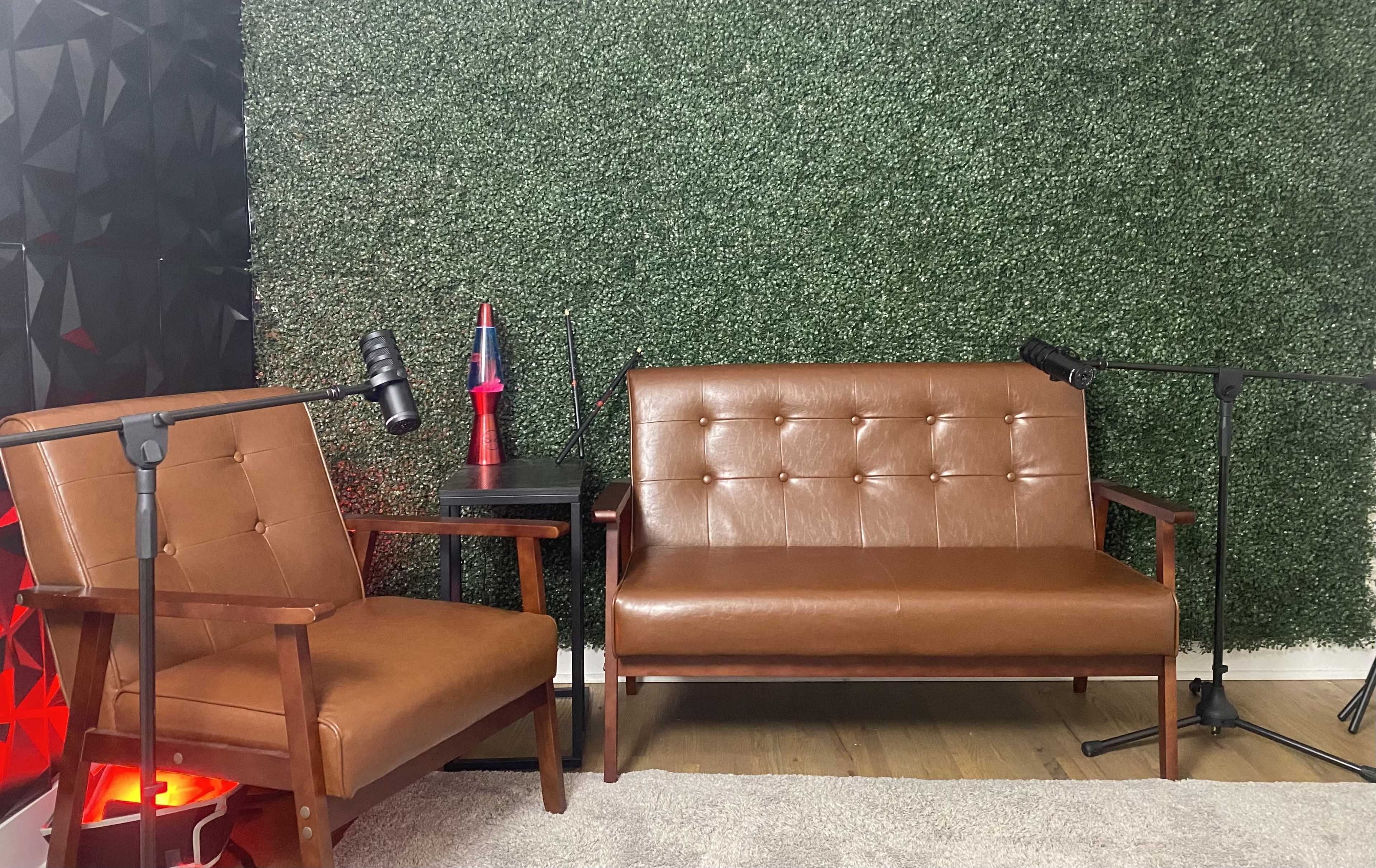 The scene shows two brown leather sofas with a small table between them, set against a green artificial hedge wall and featuring microphones on stands.