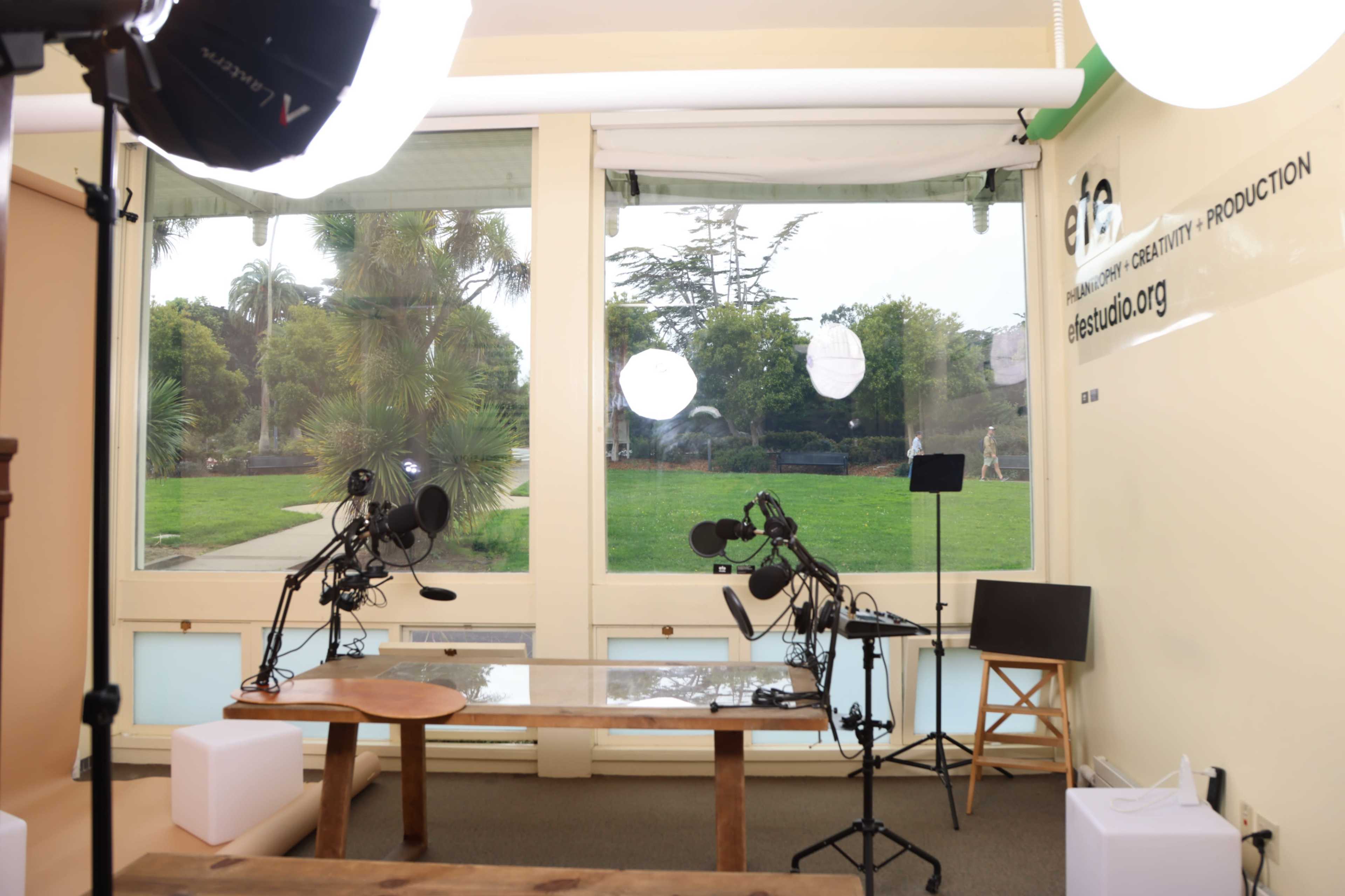 The image shows a well-lit studio space with microphones set up on stands facing a wooden table, large windows revealing a green outdoor area in the background.