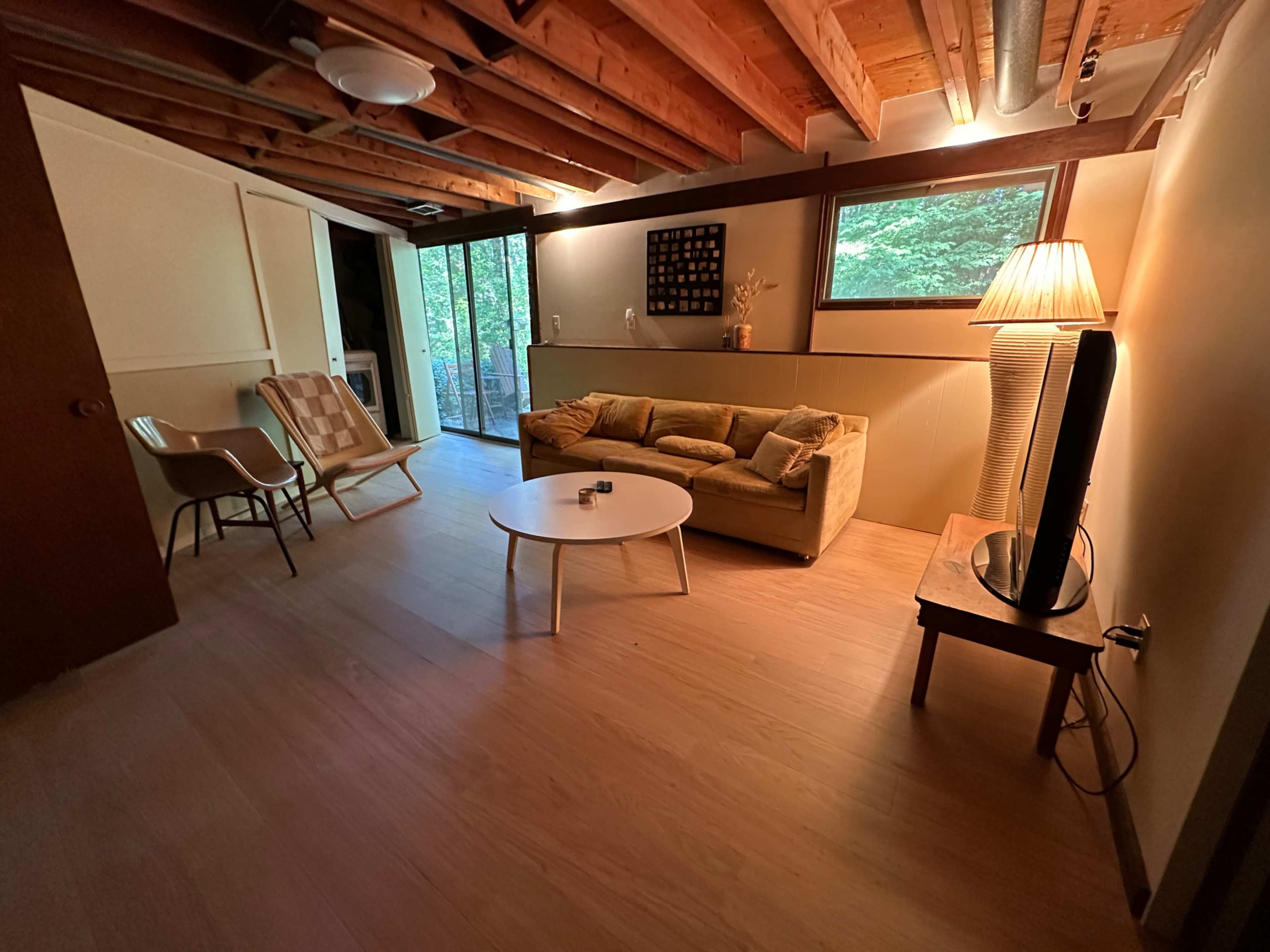The image shows a cozy living area with a sofa, a coffee table, two chairs, a floor lamp, and large windows overlooking greenery.