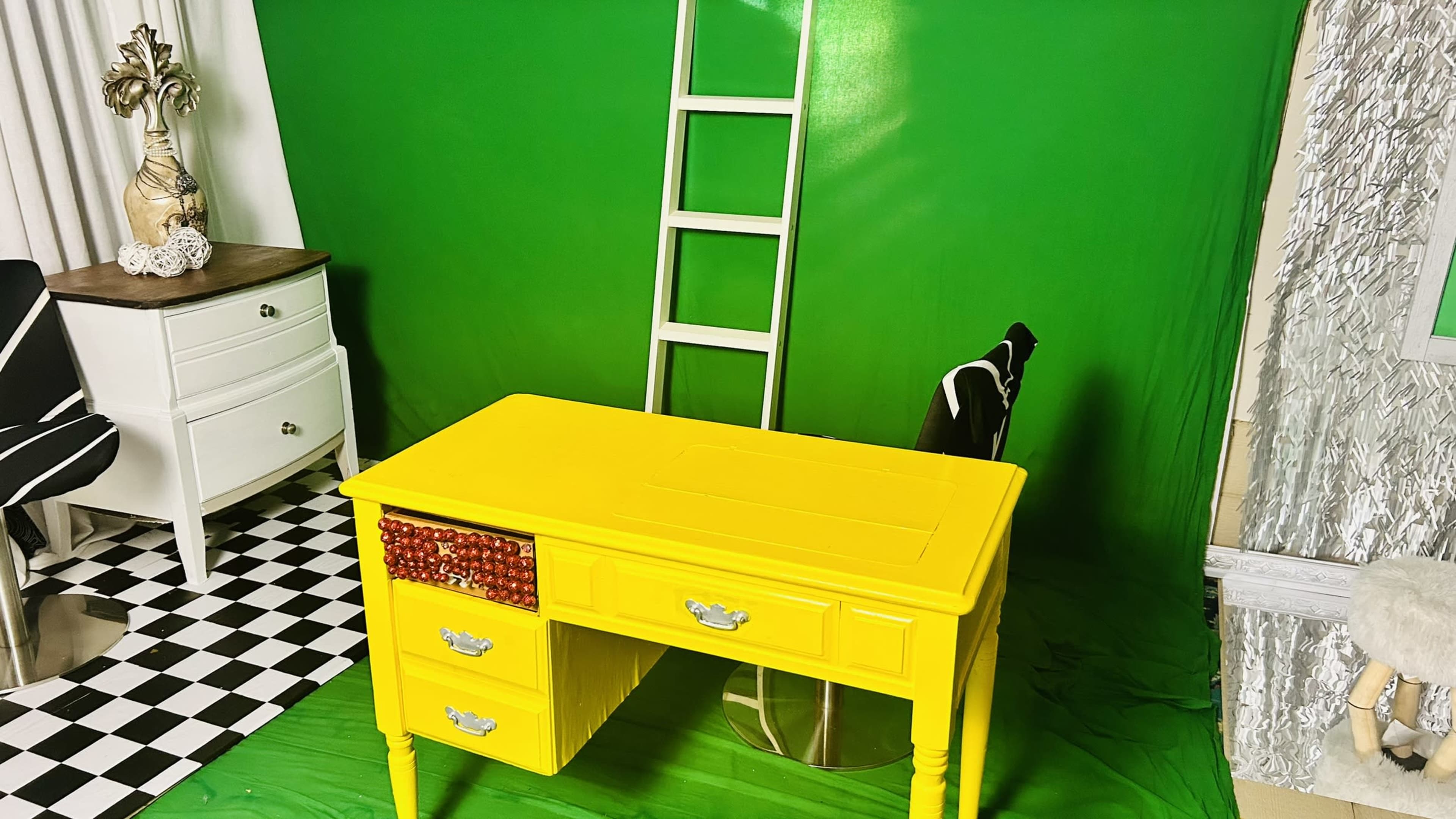 A yellow desk with drawers is positioned in front of a green backdrop, accompanied by a black and white checkered floor and a ladder.