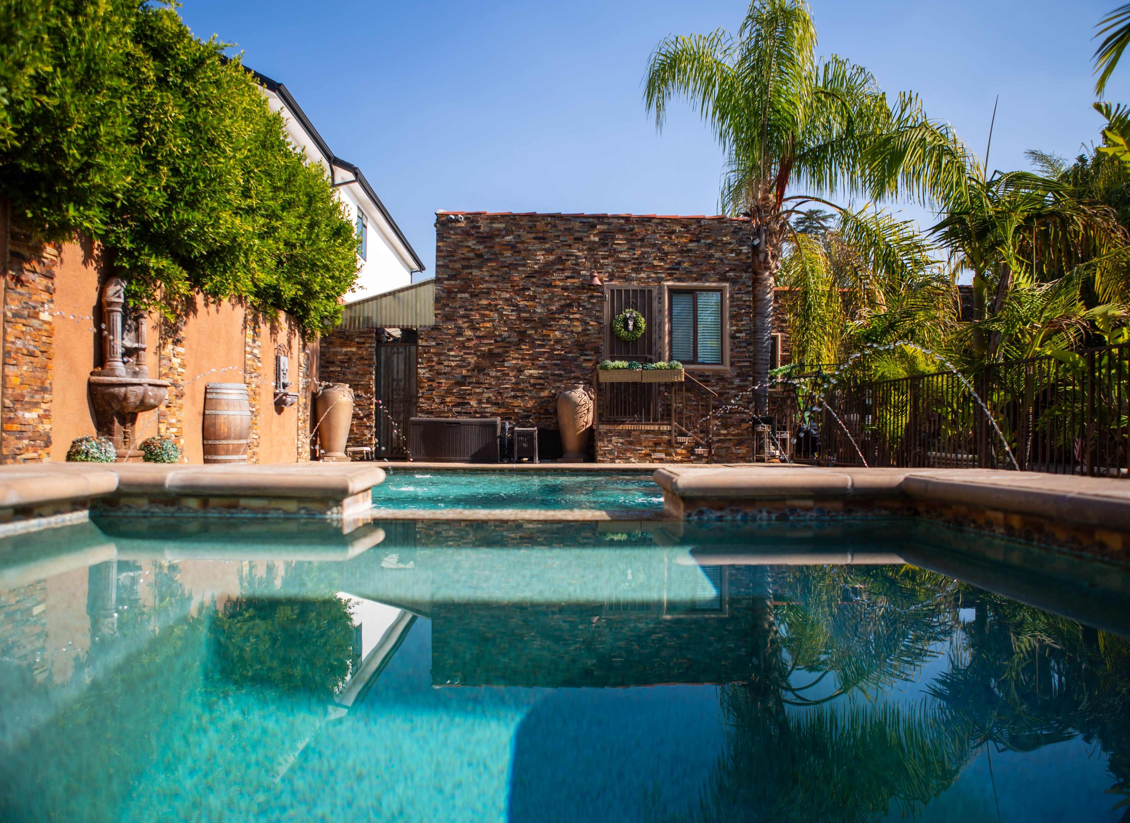 The image shows a swimming pool surrounded by tropical plants and a stone wall structure, with a clear blue sky overhead.