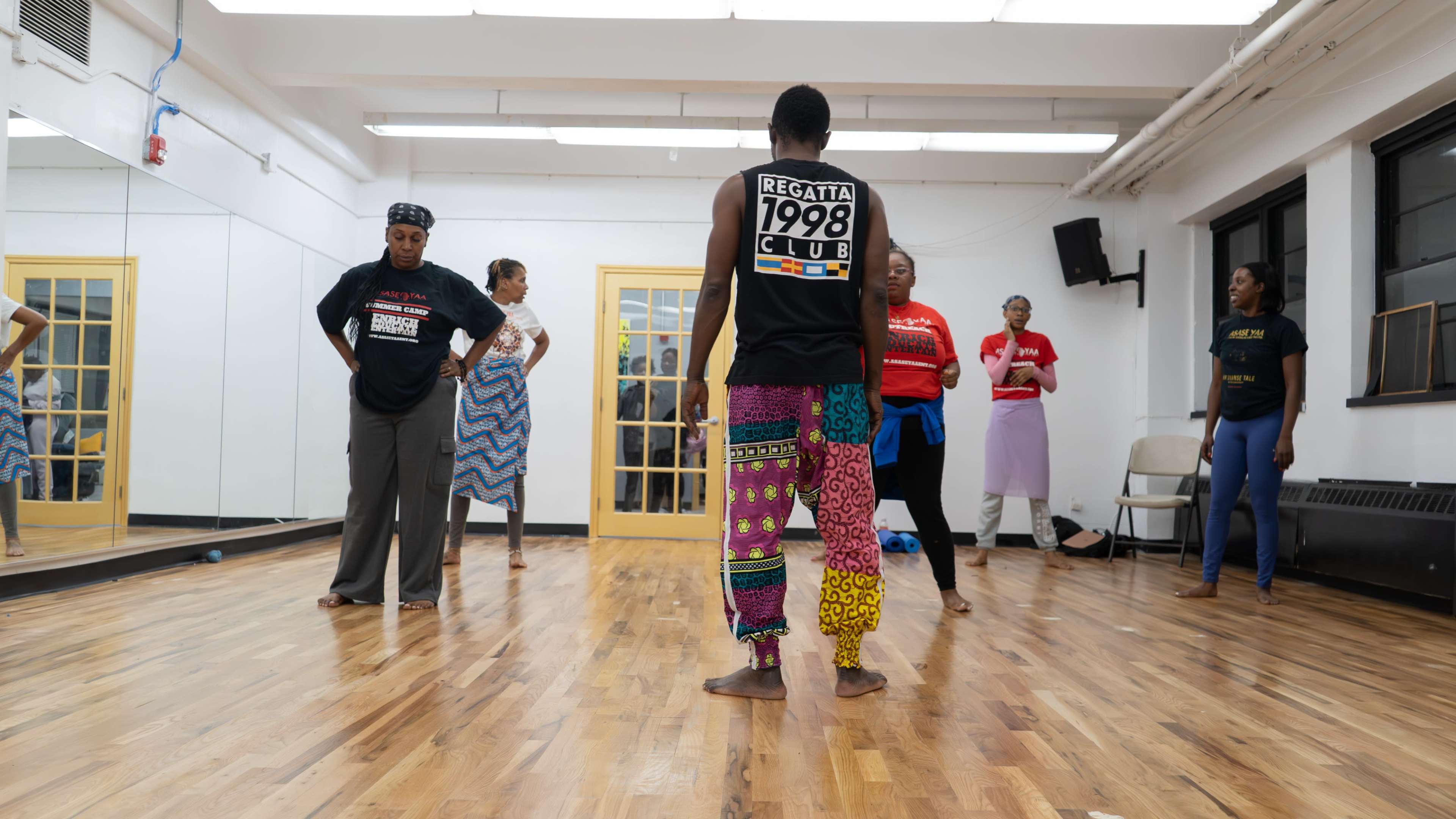 A group of dancers practices in a mirrored studio with wooden floors, showcasing a variety of clothing styles.