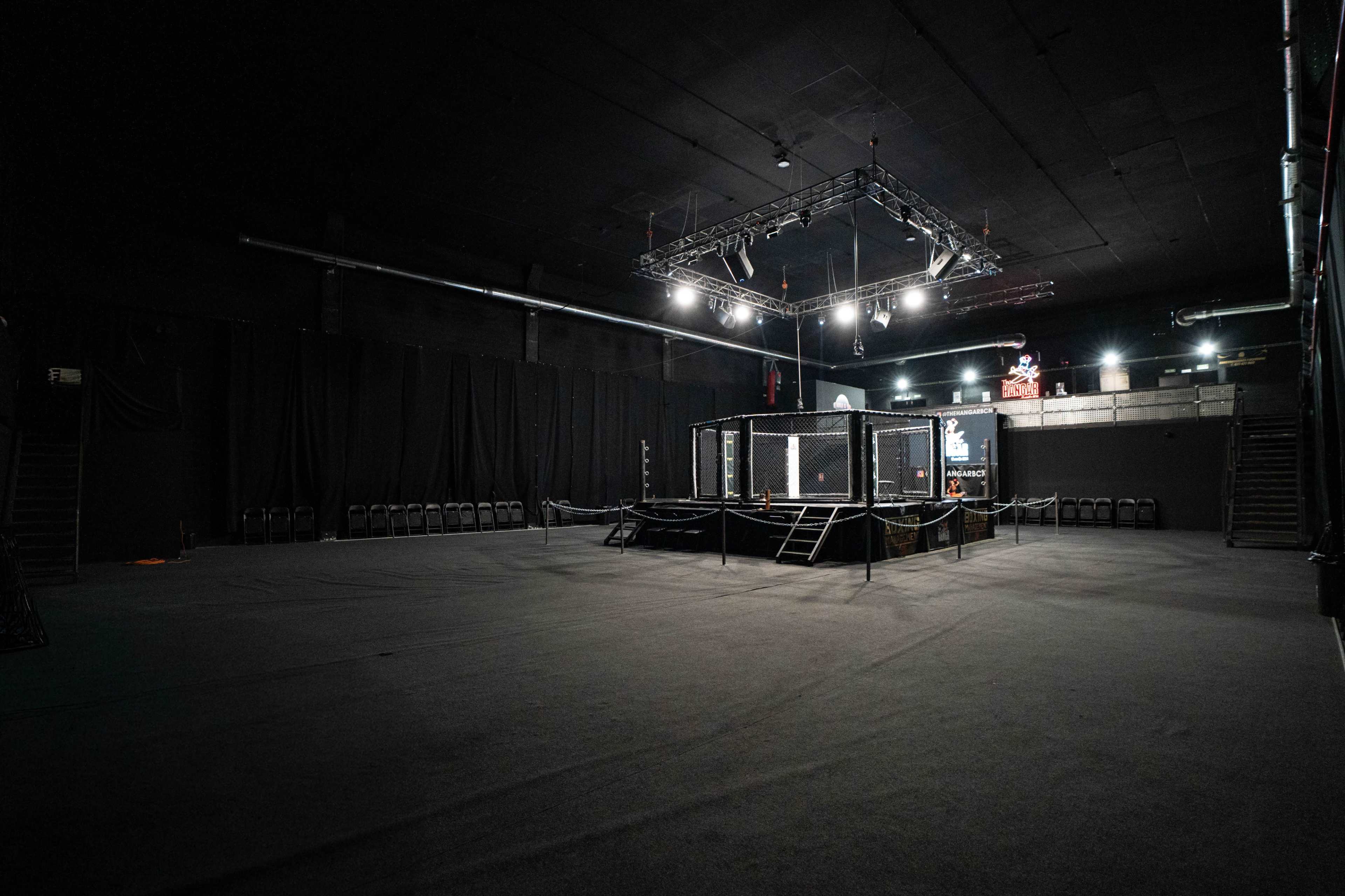 An empty boxing ring enclosed by a cage is situated in a dimly lit arena with black walls and minimal seating.