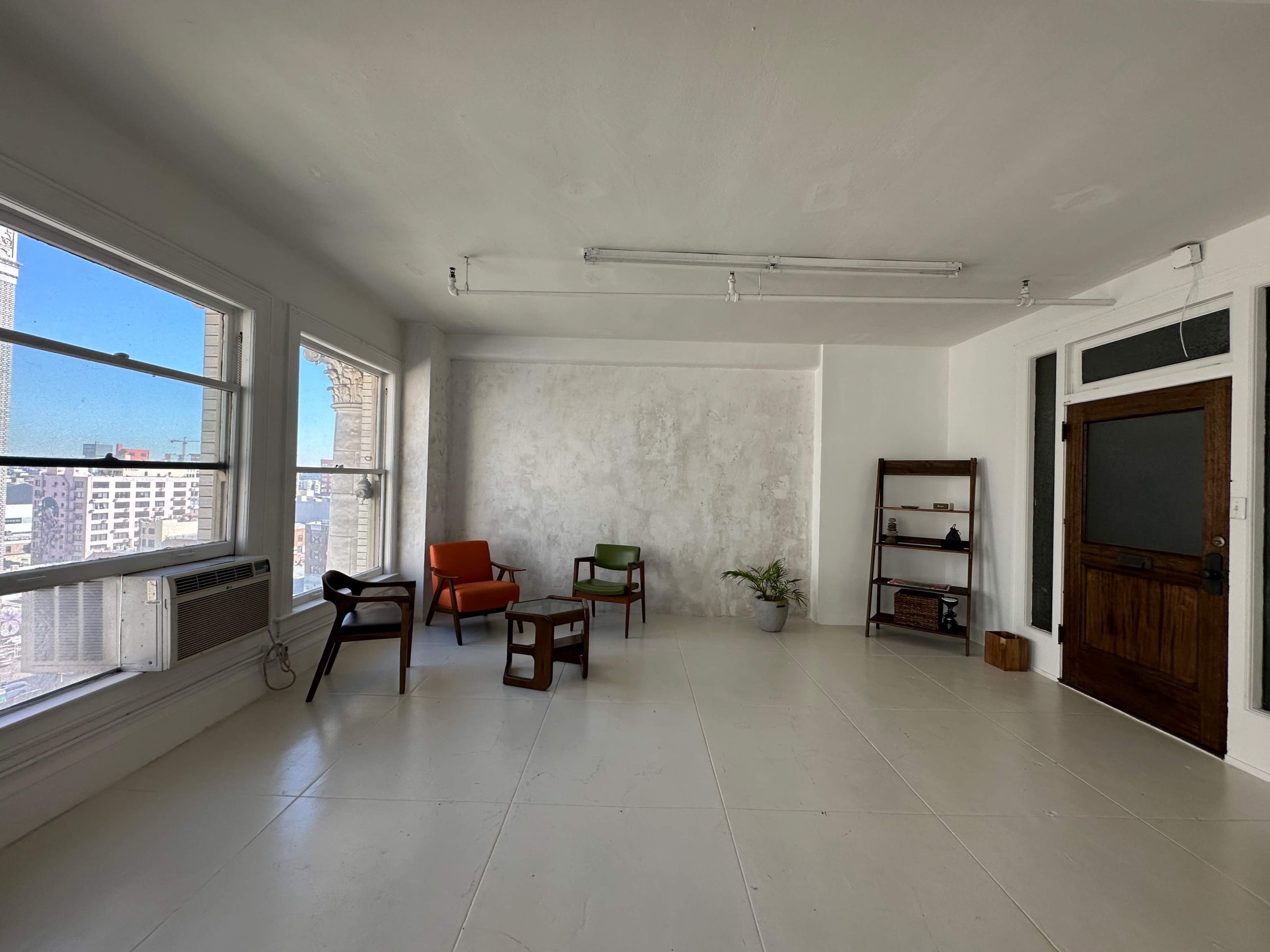 A spacious, empty room with large windows, featuring a few pieces of furniture, including two armchairs, a coffee table, and a shelf.