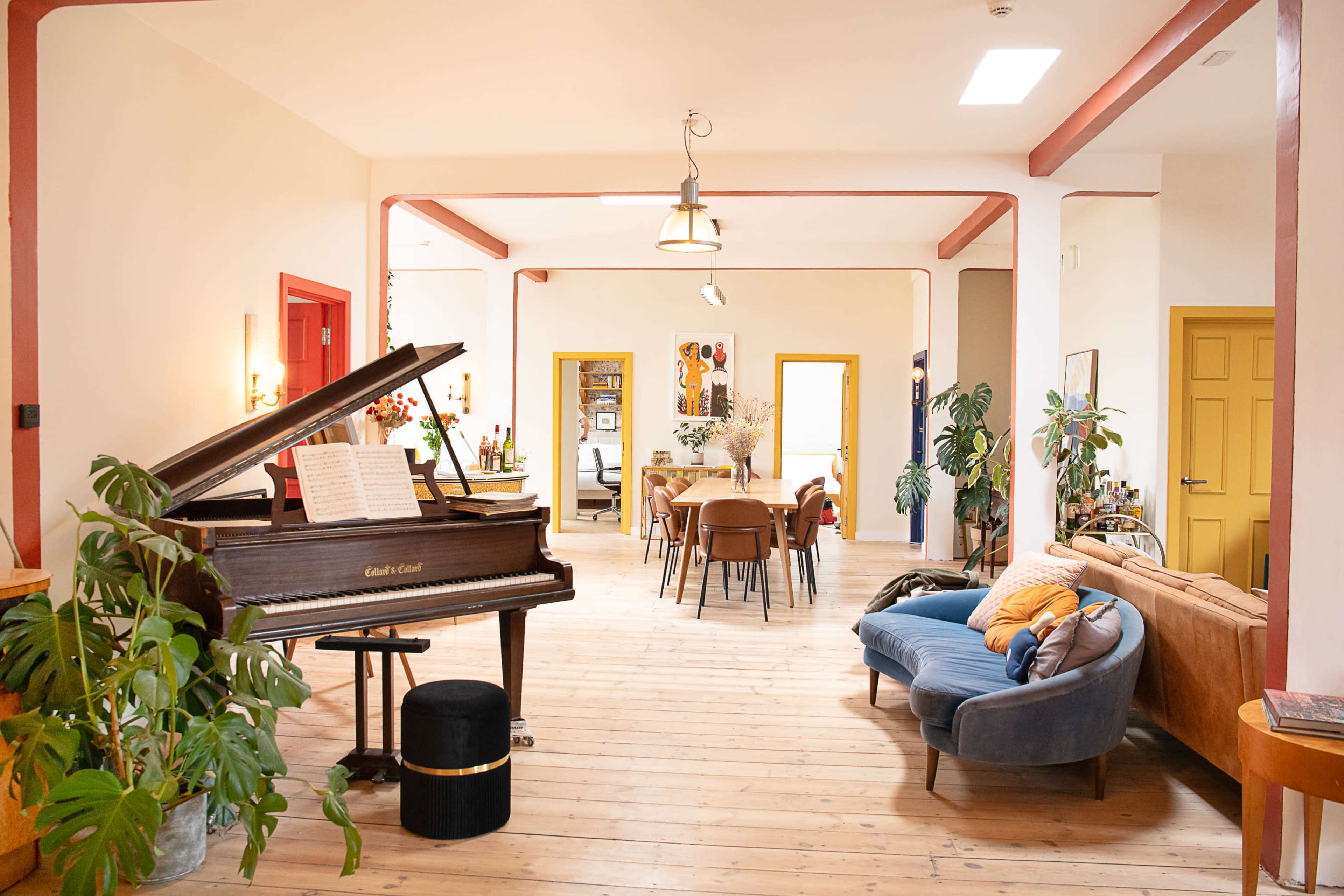 The image shows a spacious, well-lit room featuring a grand piano, seating area with a blue sofa, and a dining space with multiple chairs.