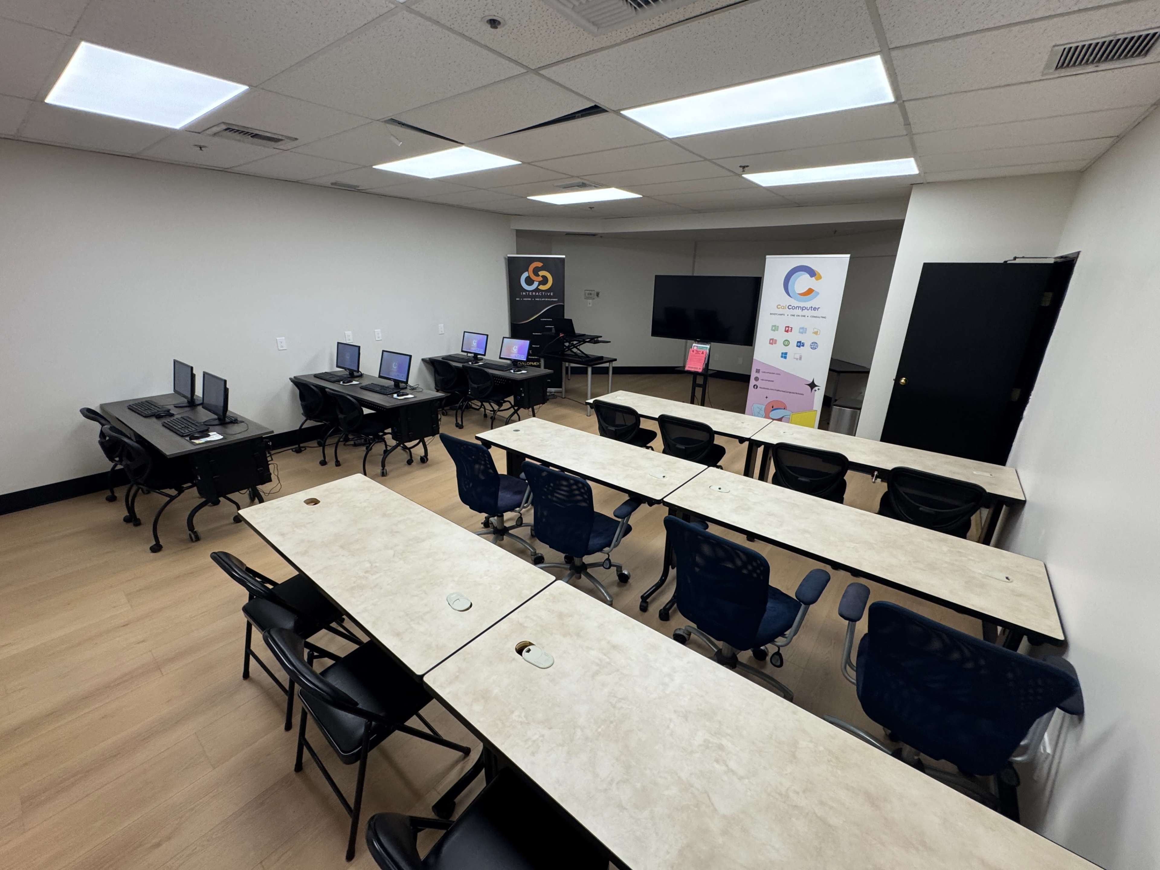 The image shows a spacious classroom with several desks and chairs arranged in rows, two groups of computer stations, and a large screen at the front of the room.