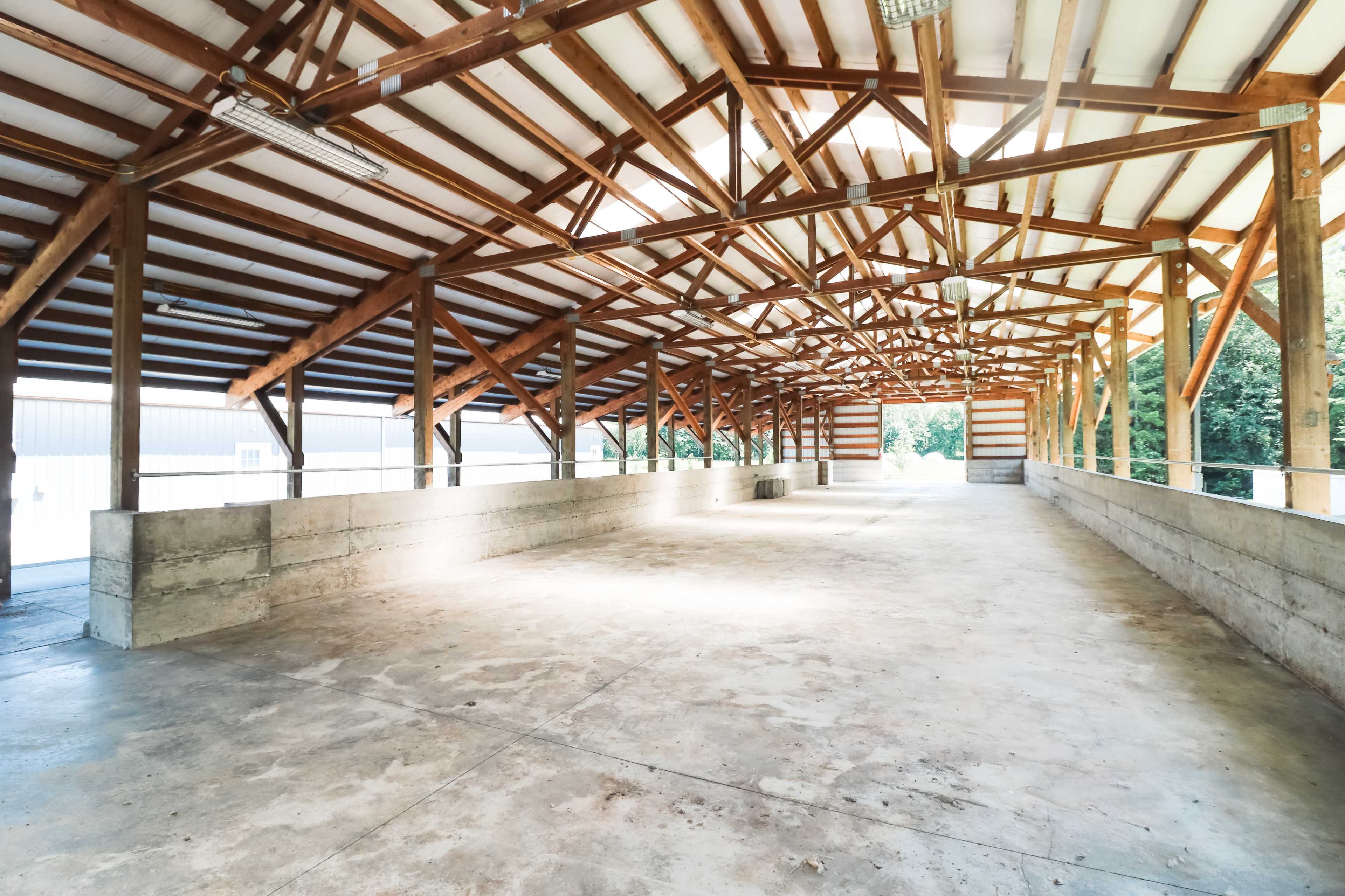 The image shows a spacious indoor arena with wooden beams and a concrete floor, illuminated by natural light.