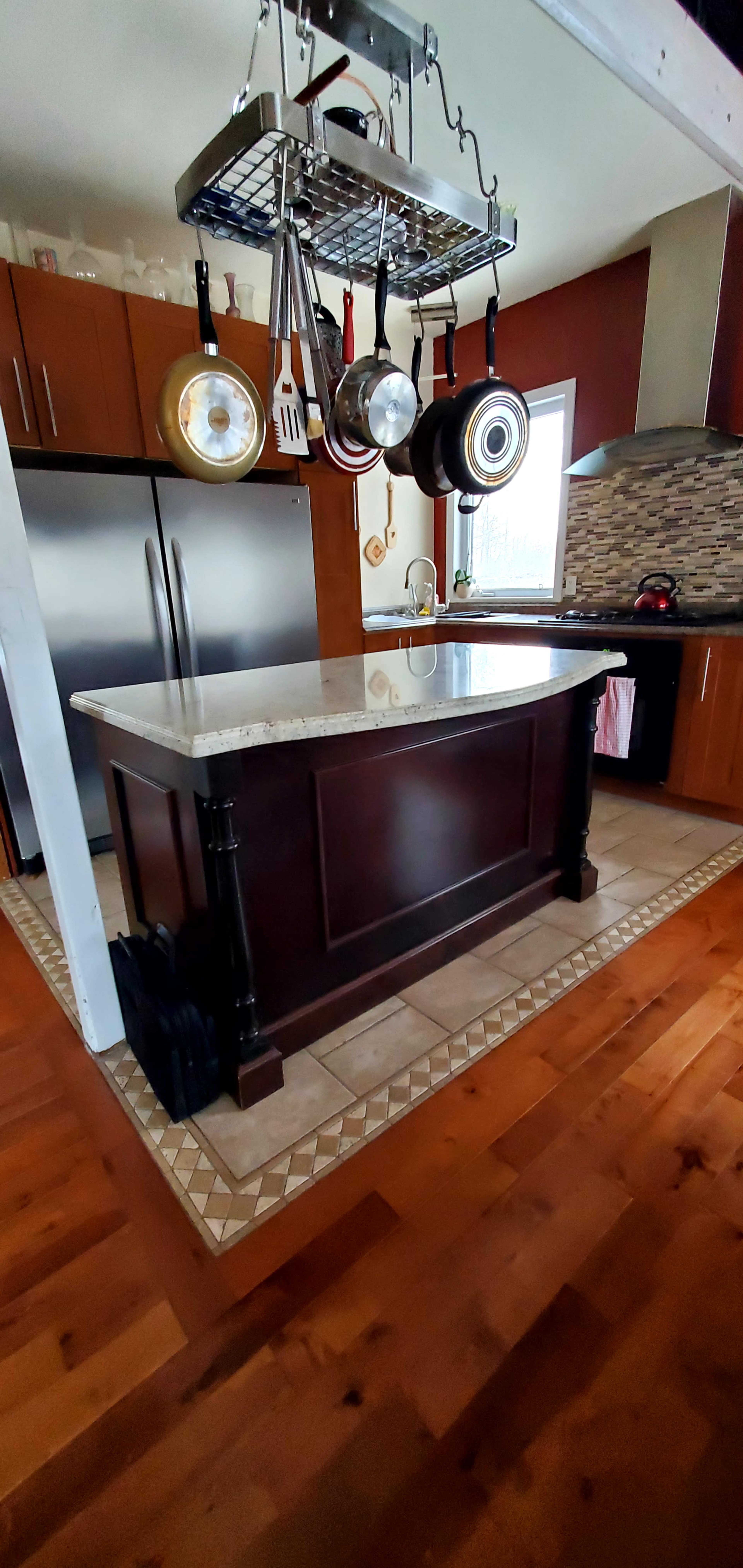 A kitchen features a central island with a granite countertop and a hanging pot rack above it, surrounded by wooden cabinets and a tiled floor.