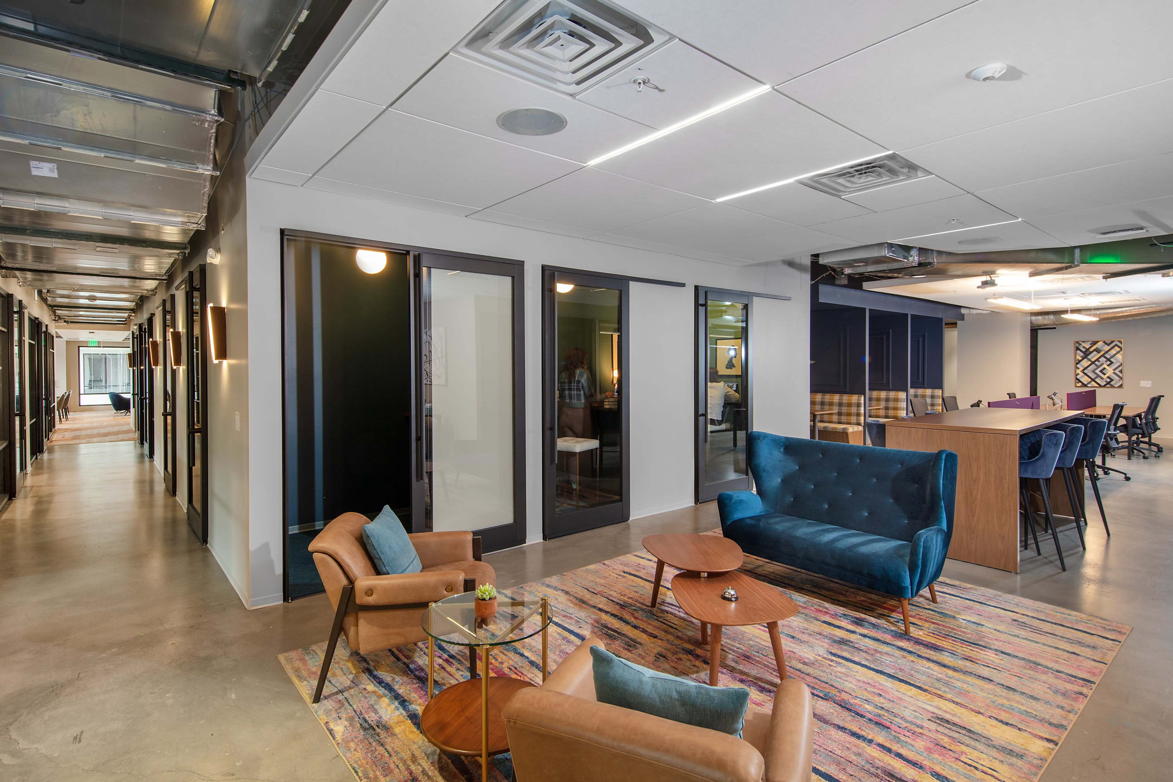 The image shows a modern office space featuring a lounge area with a blue sofa and two chairs, surrounded by glass-walled meeting rooms and a communal workspace.