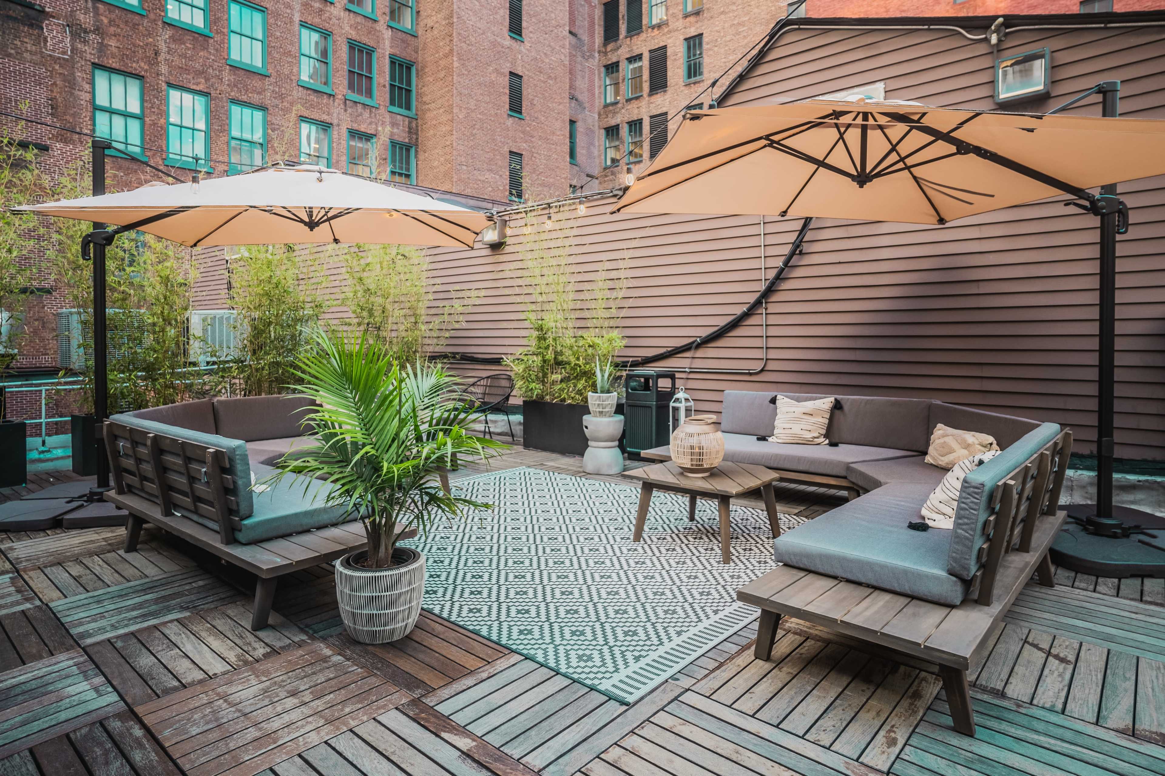 The image shows a rooftop terrace furnished with lounge seating, large umbrellas, and potted plants, surrounded by brick buildings.