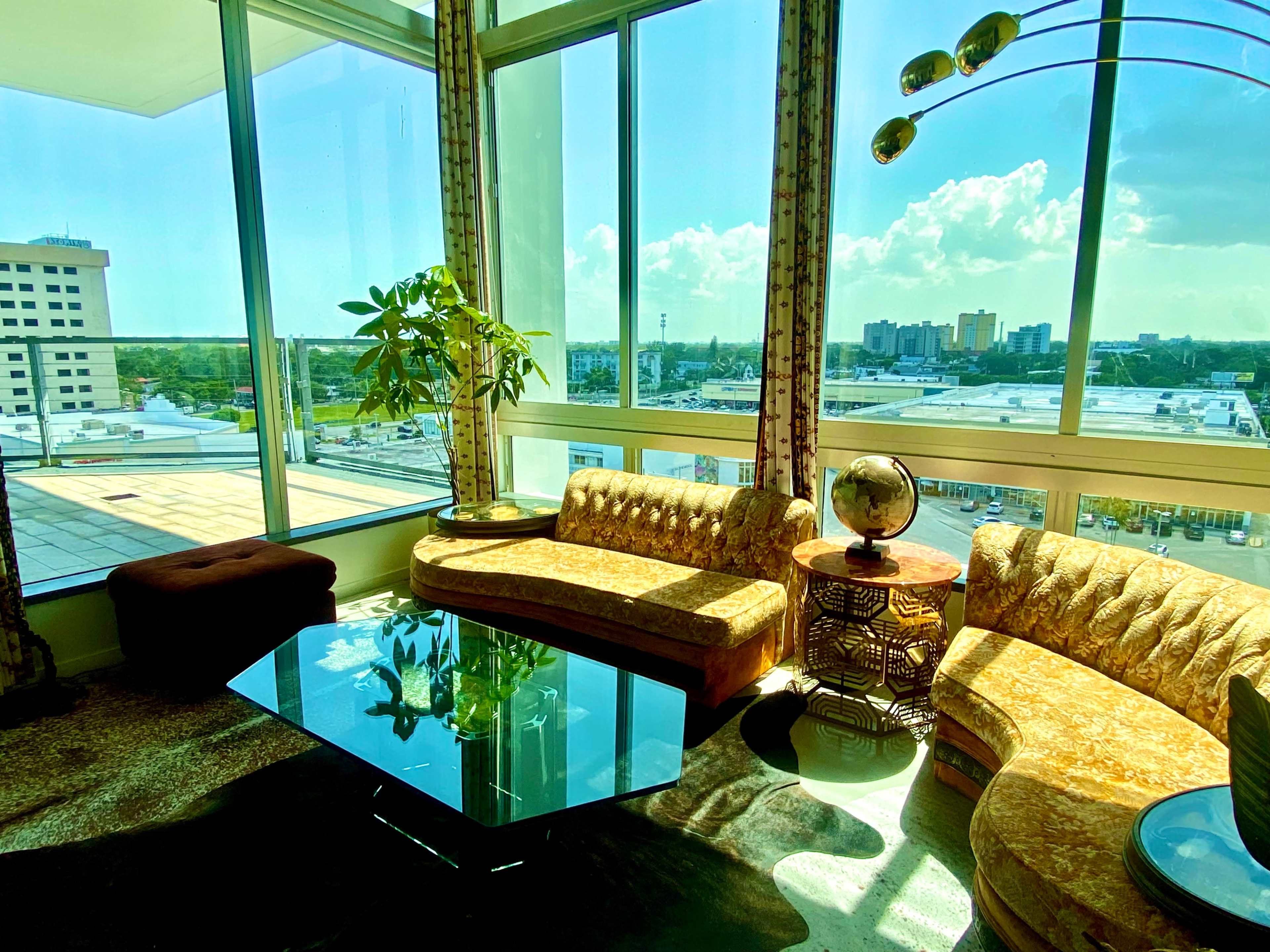 The image shows a modern living room with a curved golden-brown sofa, a glass coffee table, and large windows offering a view of a city skyline.