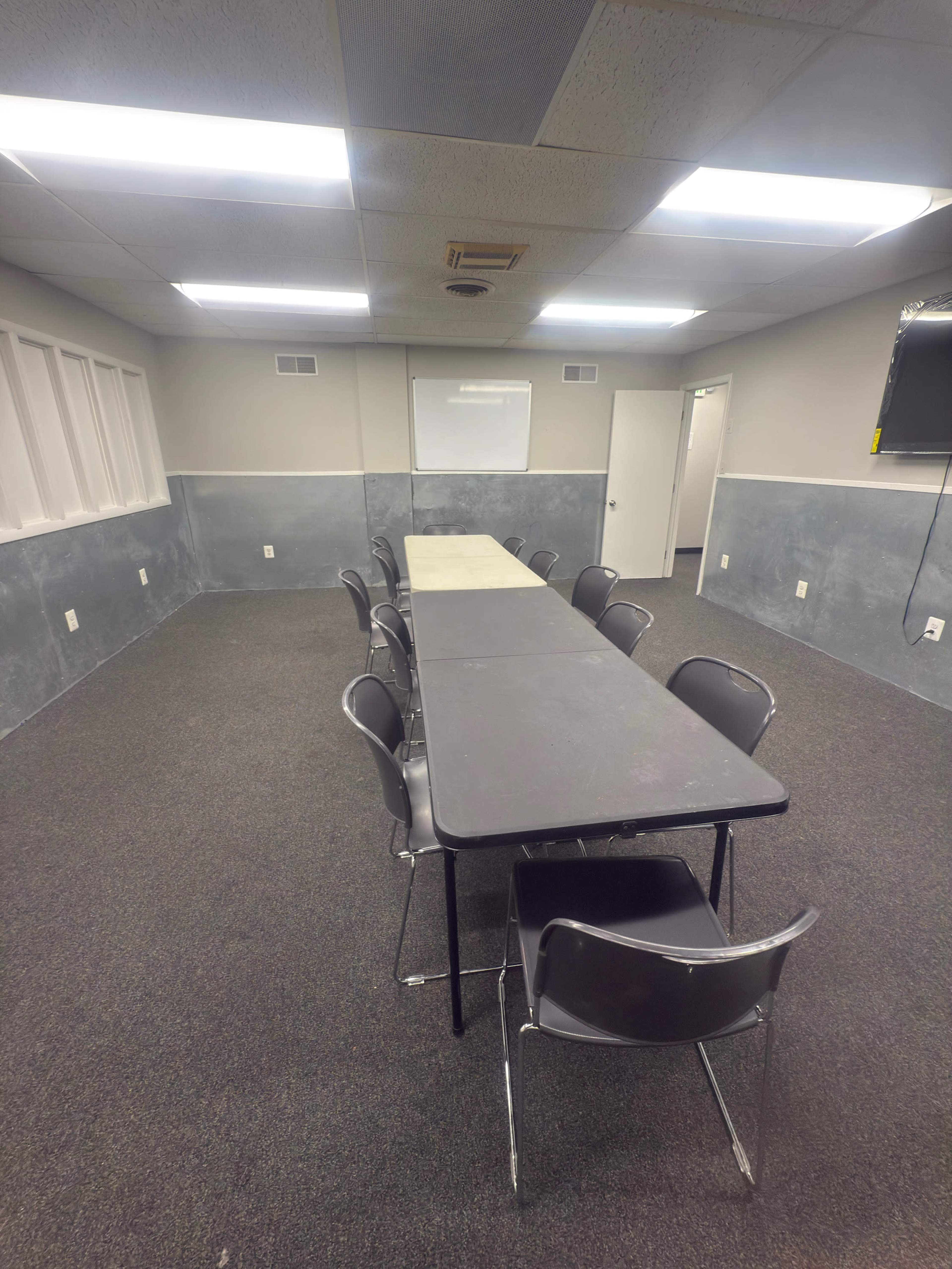 The image shows a sparsely furnished meeting room with a long table and several chairs arranged around it, featuring gray walls and ceiling lights.