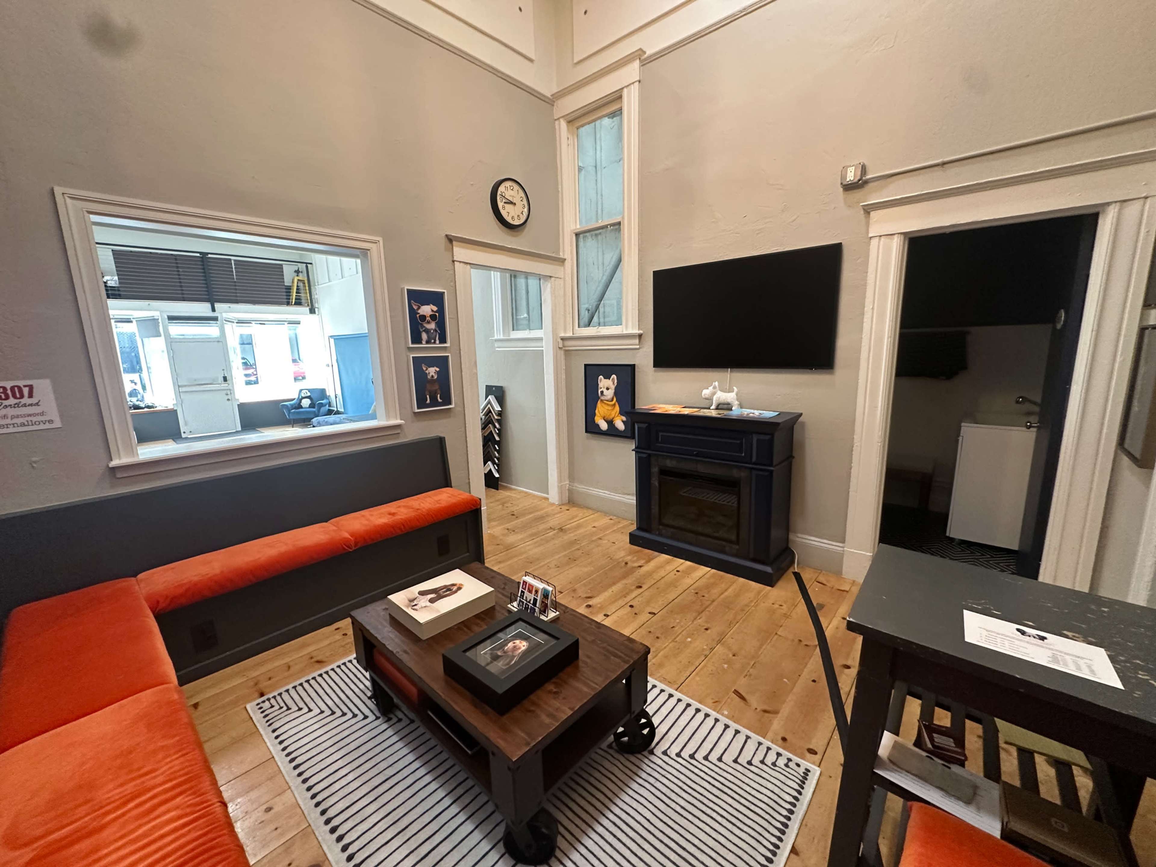 The image shows a cozy interior of a room featuring a gray wall, an orange sofa, a fireplace, a television mounted on the wall, and a small dining area with a table.