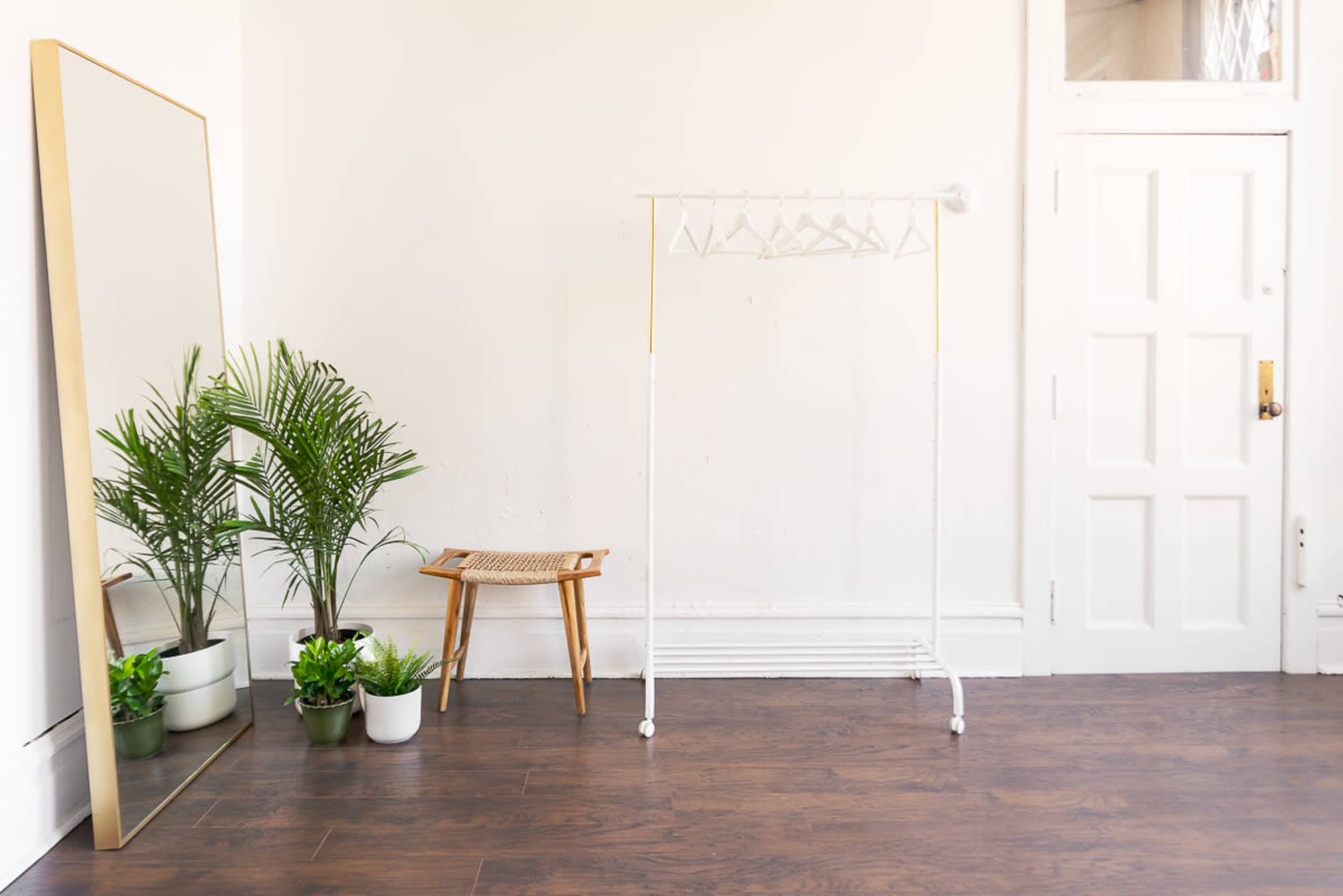 A minimalist room featuring a clothing rack, a large mirror, a small stool, and several potted plants against a white wall.