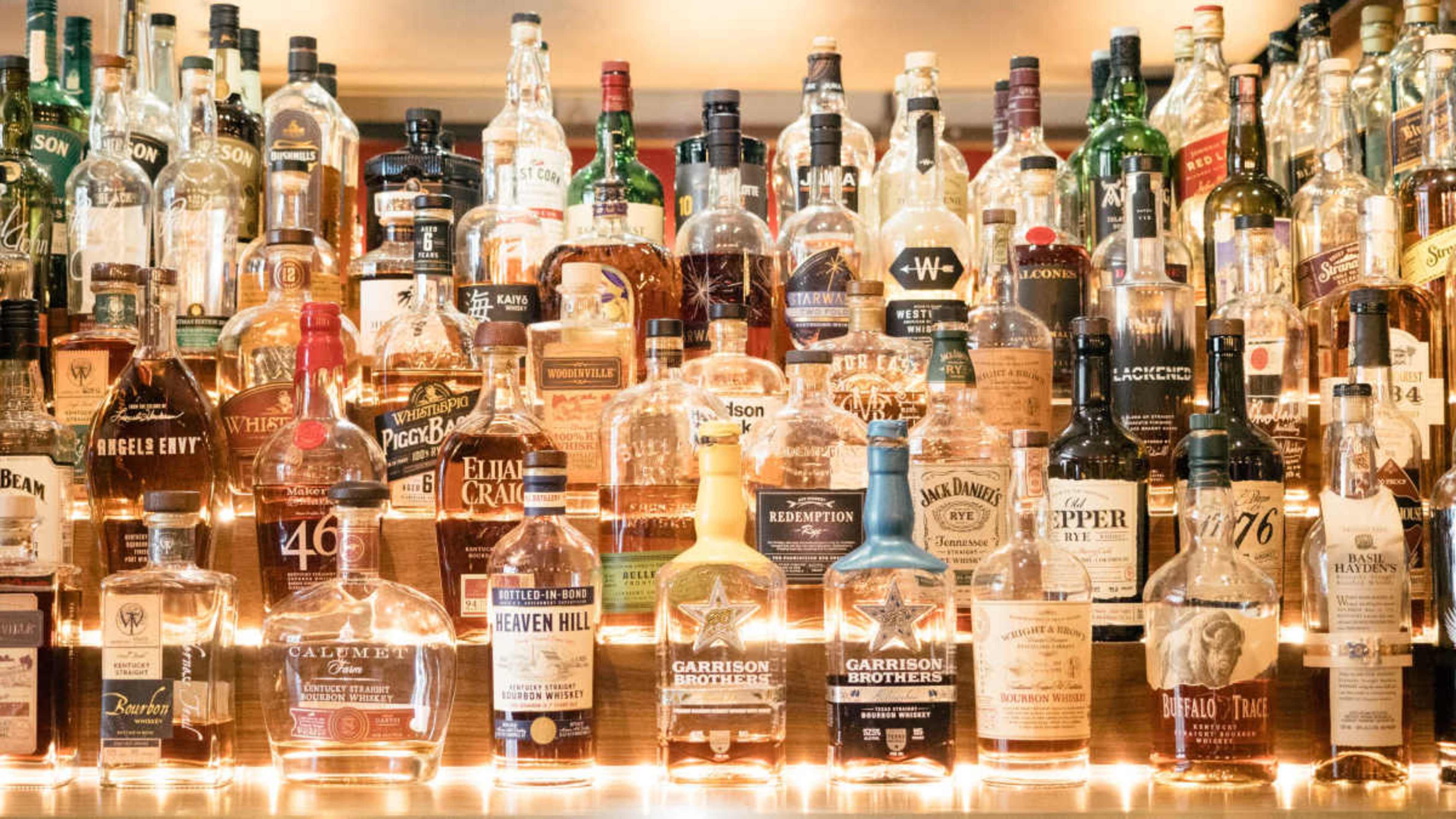 A bar shelf displays a wide array of bottles filled with various types of spirits, arranged neatly with some illuminated by soft lighting.