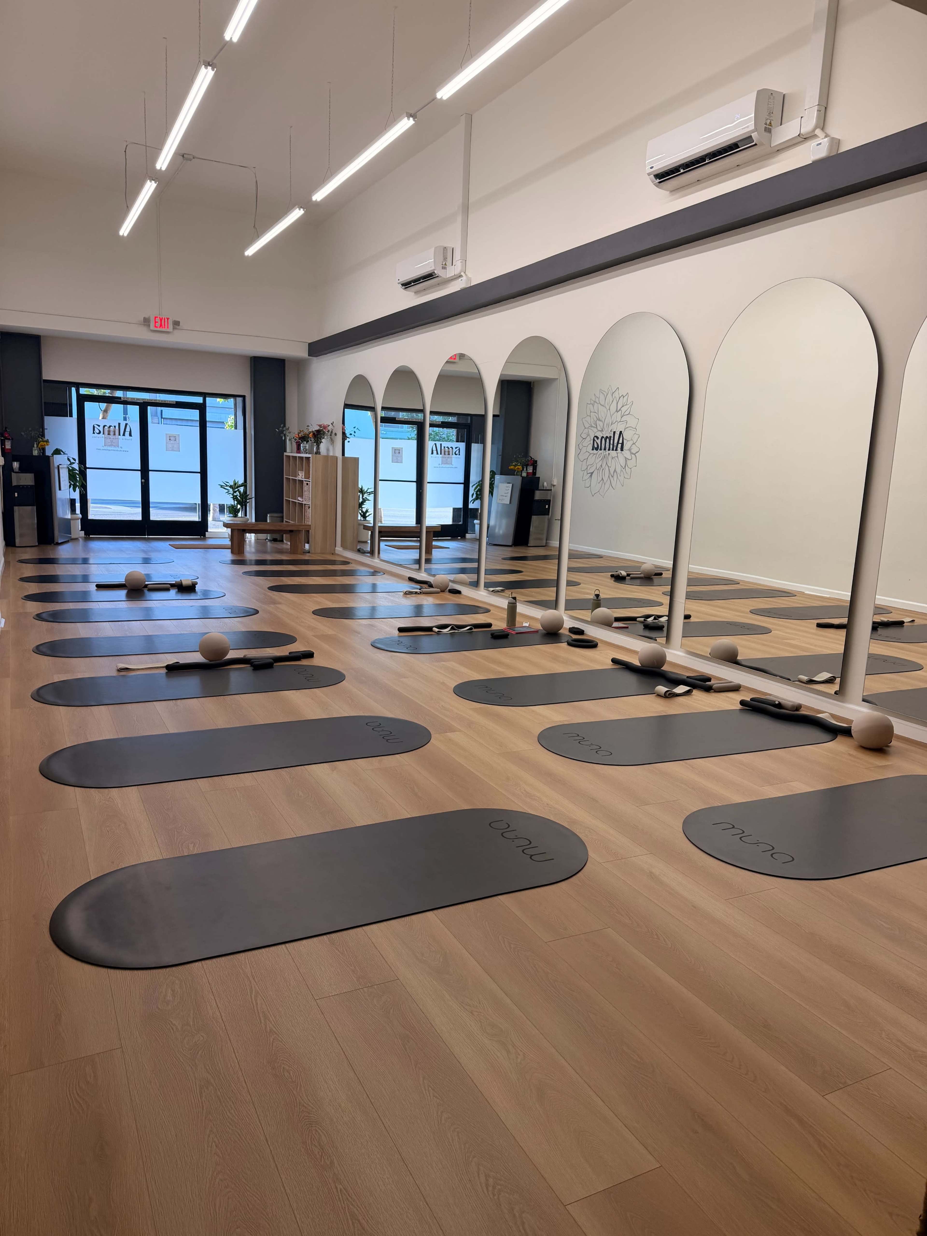 The image shows a yoga studio with several exercise mats arranged on a wooden floor, accompanied by large mirrors and a welcoming entrance.