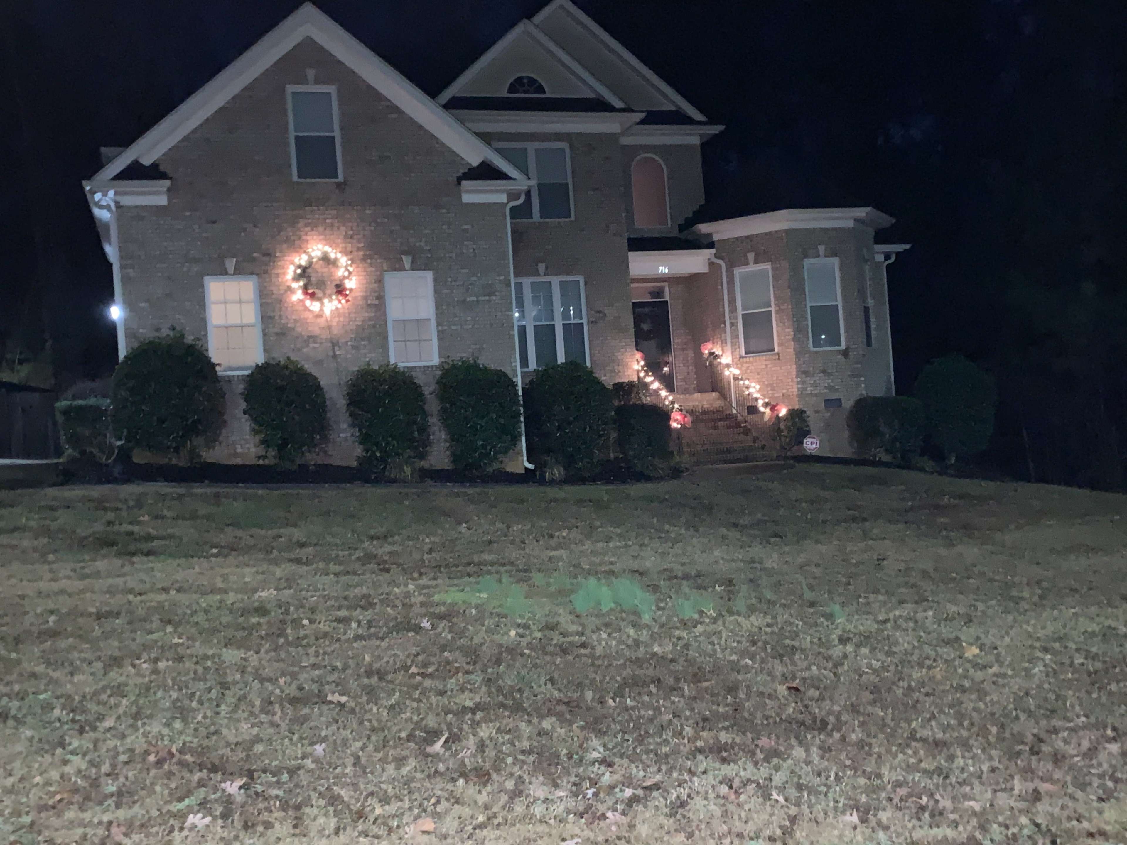 A two-story brick house is illuminated by decorative lights and wreaths, set on a grassy lawn at night.