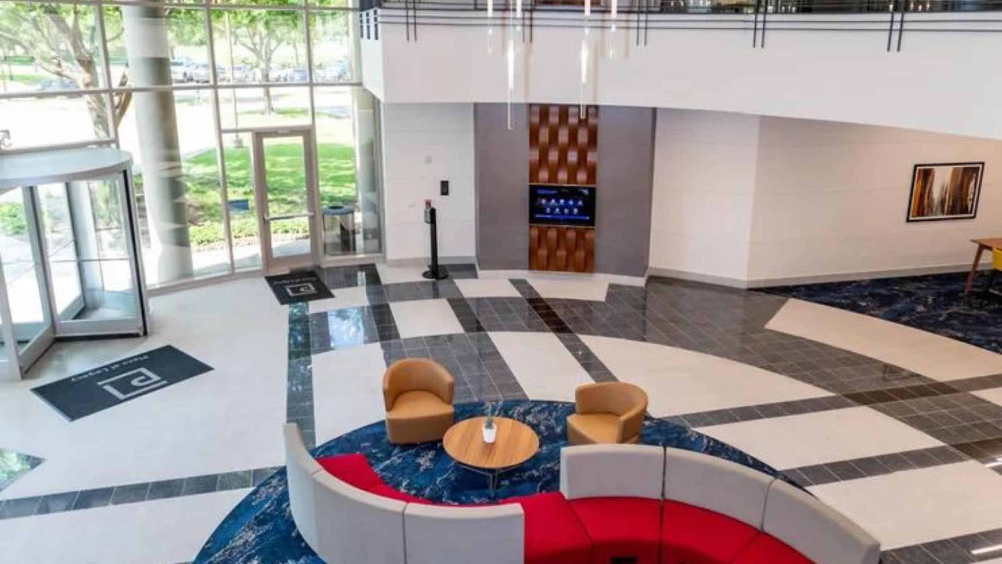The image shows a modern lobby with a circular seating area, a coffee table, and large windows allowing natural light to enter.