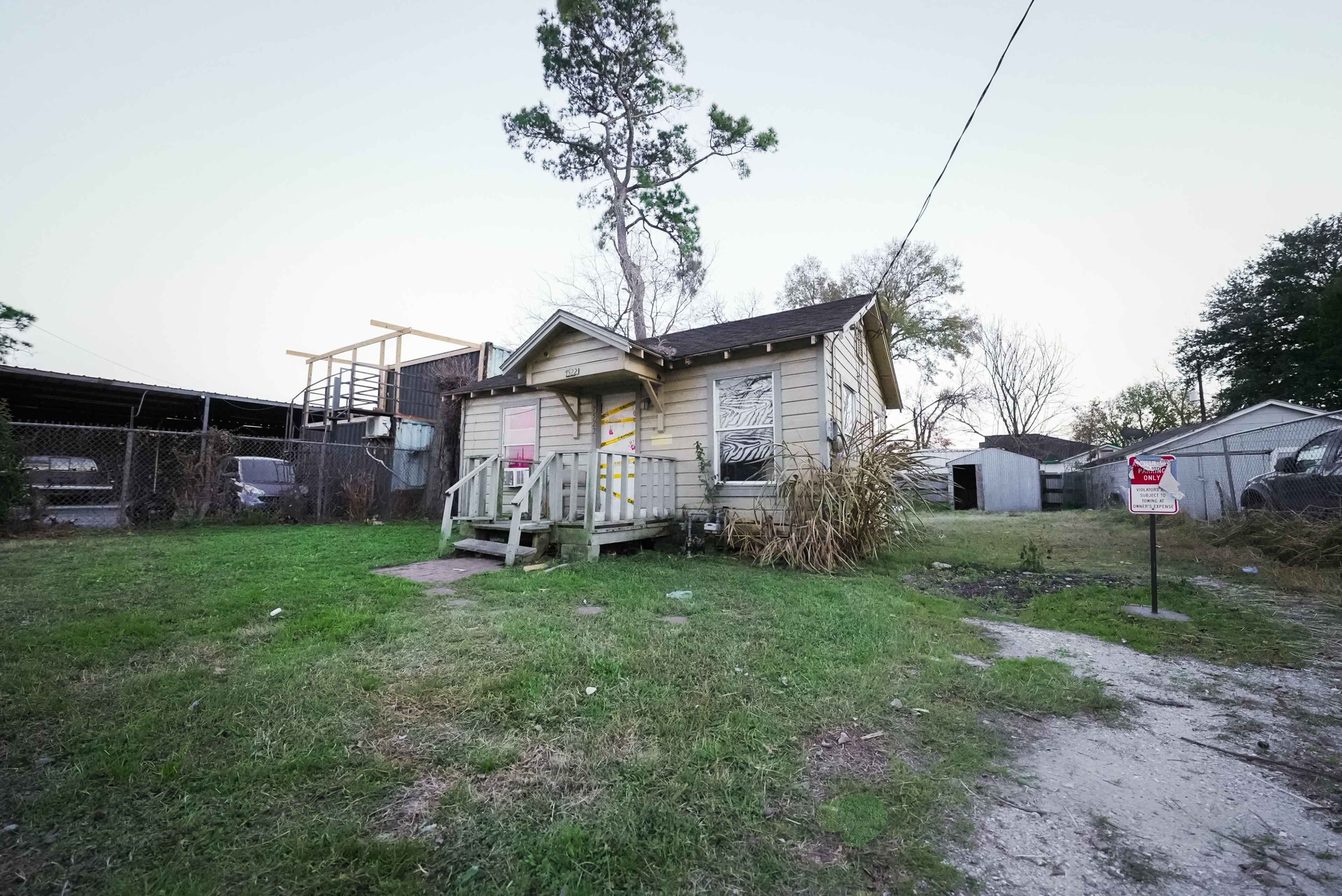 A small, weathered house with a front porch is situated on a grassy lot, surrounded by other structures and trees.