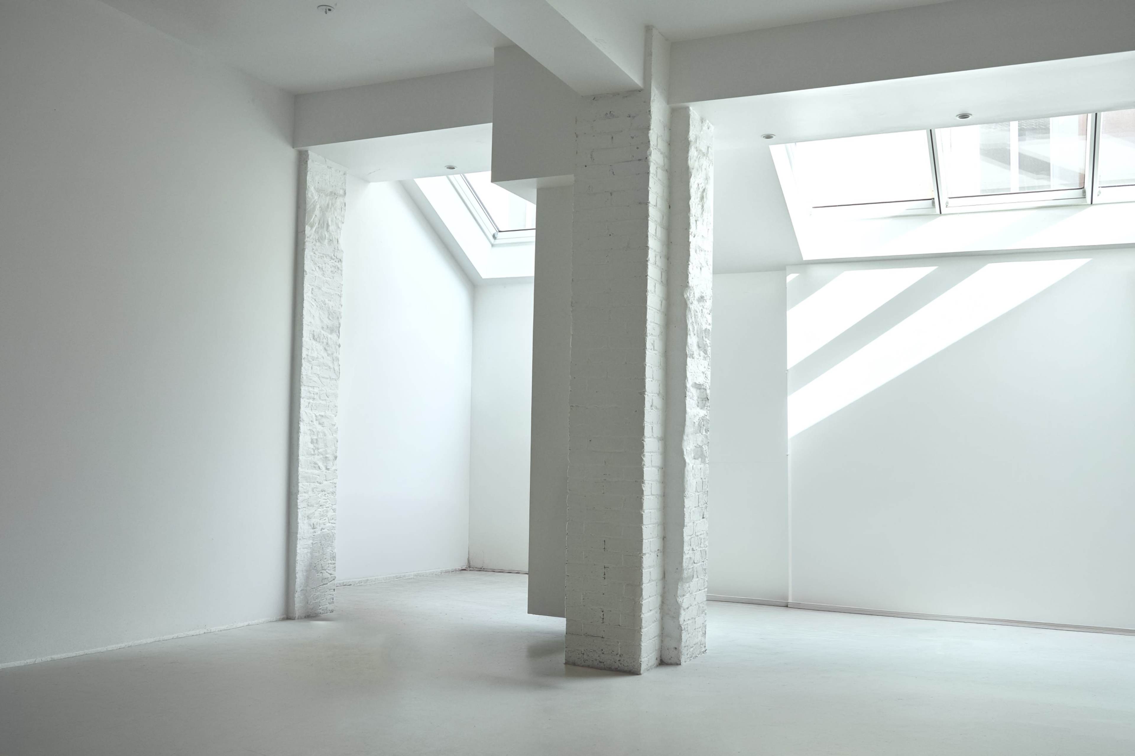 The image shows a minimalistic interior space with white walls, tall windows allowing natural light, and exposed columns.