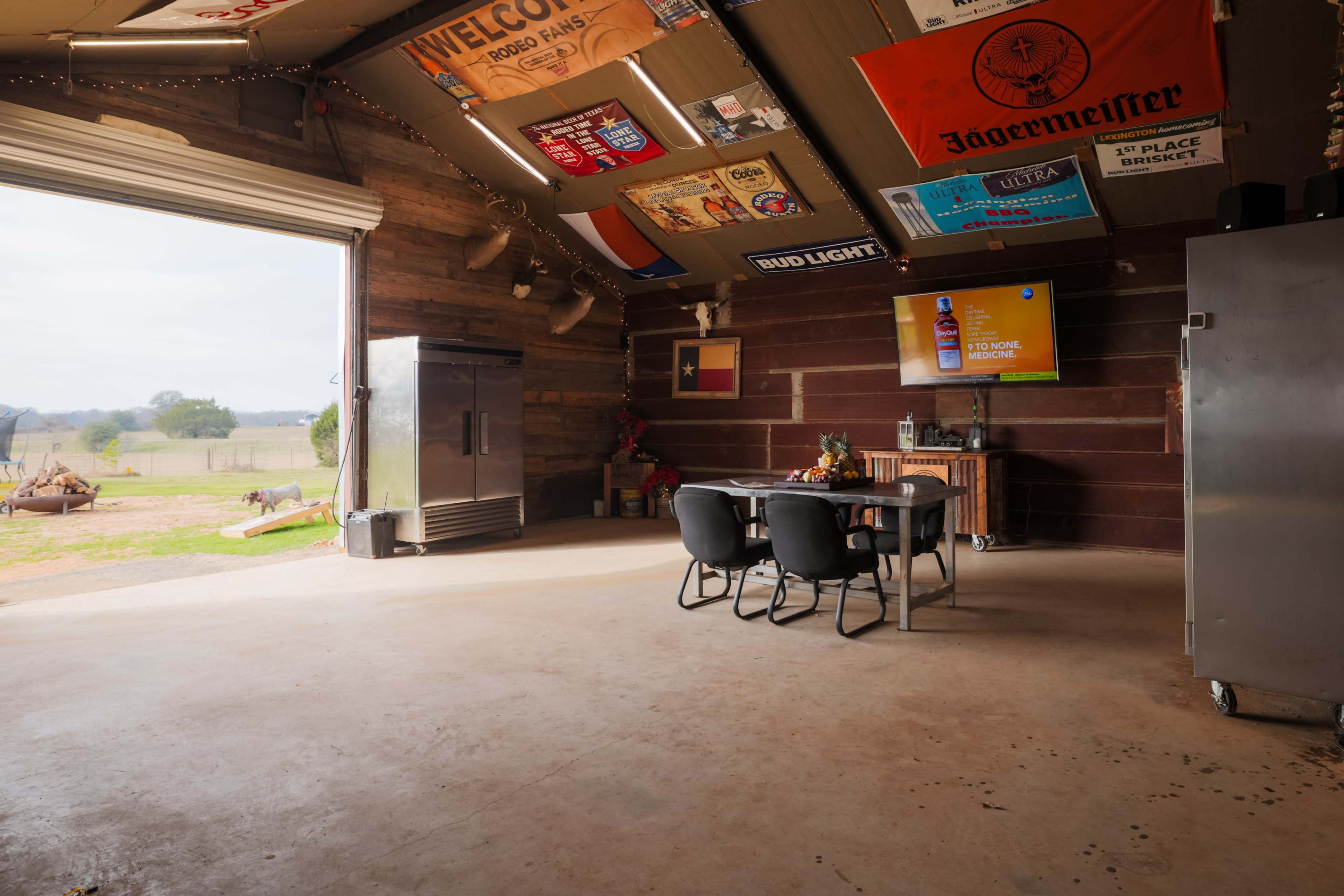 A table with four chairs sits in a rustic barn, which features flags hanging from the ceiling and a large door open to a view of the landscape outside.