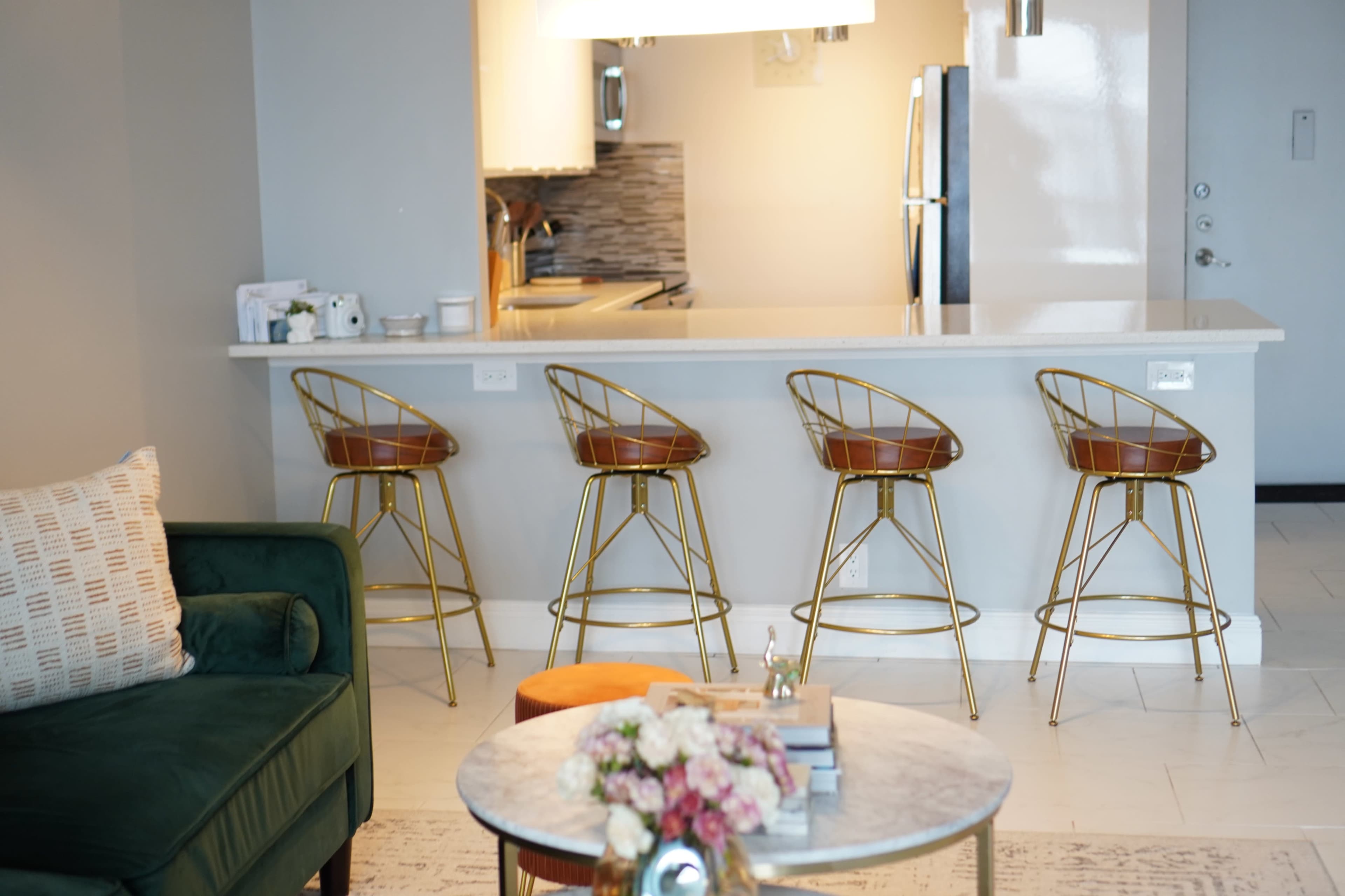 A modern kitchen bar area features four gold-accented stools lined up at a white countertop, with a glimpse of kitchen appliances and decor in the background.