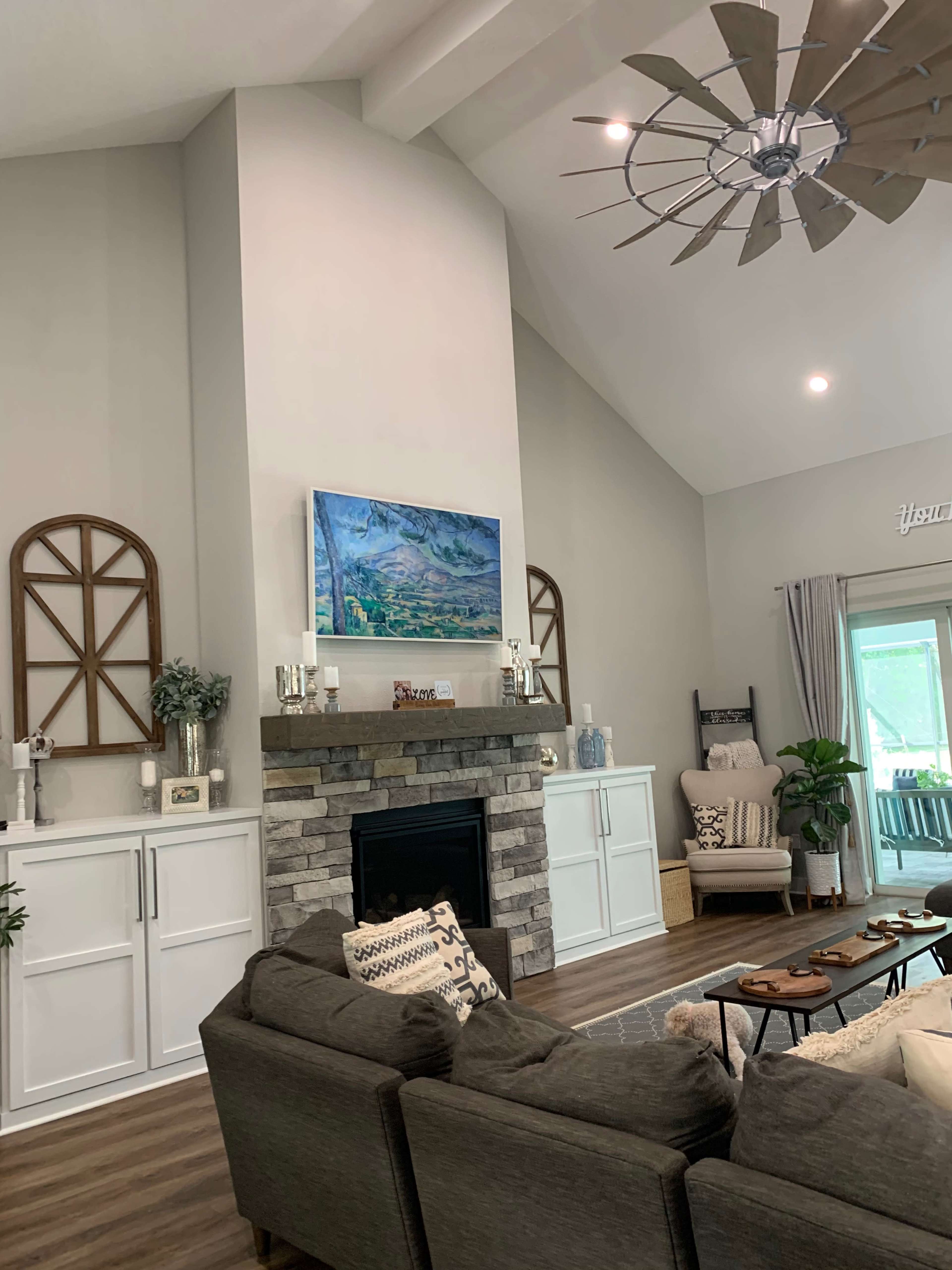 The image shows a living room with a tall ceiling, featuring a stone fireplace beneath a mounted television, flanked by decorative wooden accents and modern furniture.