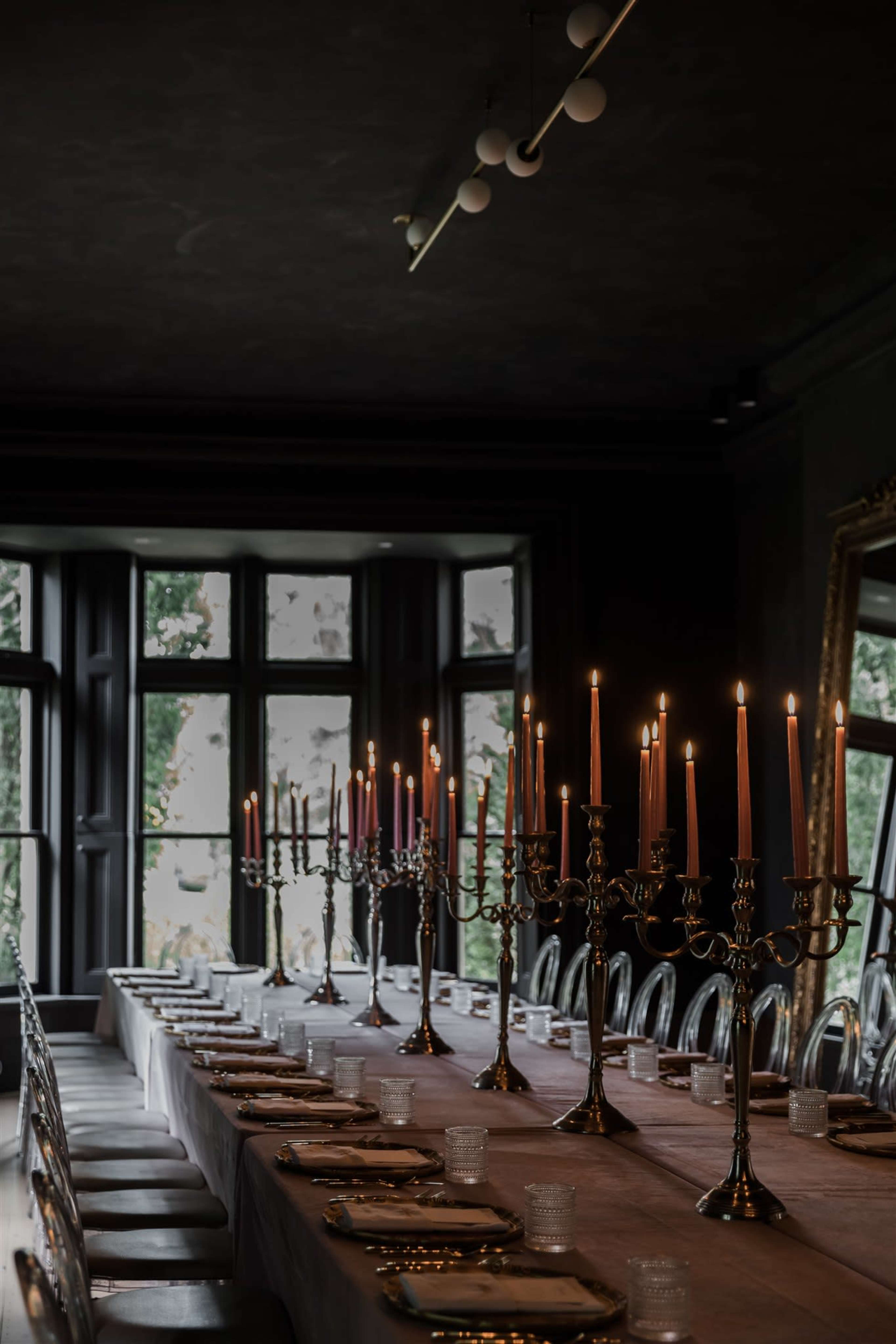 A long dining table is set with candelabras and plates, illuminated by the glow of candles, in a dimly lit room with large windows.