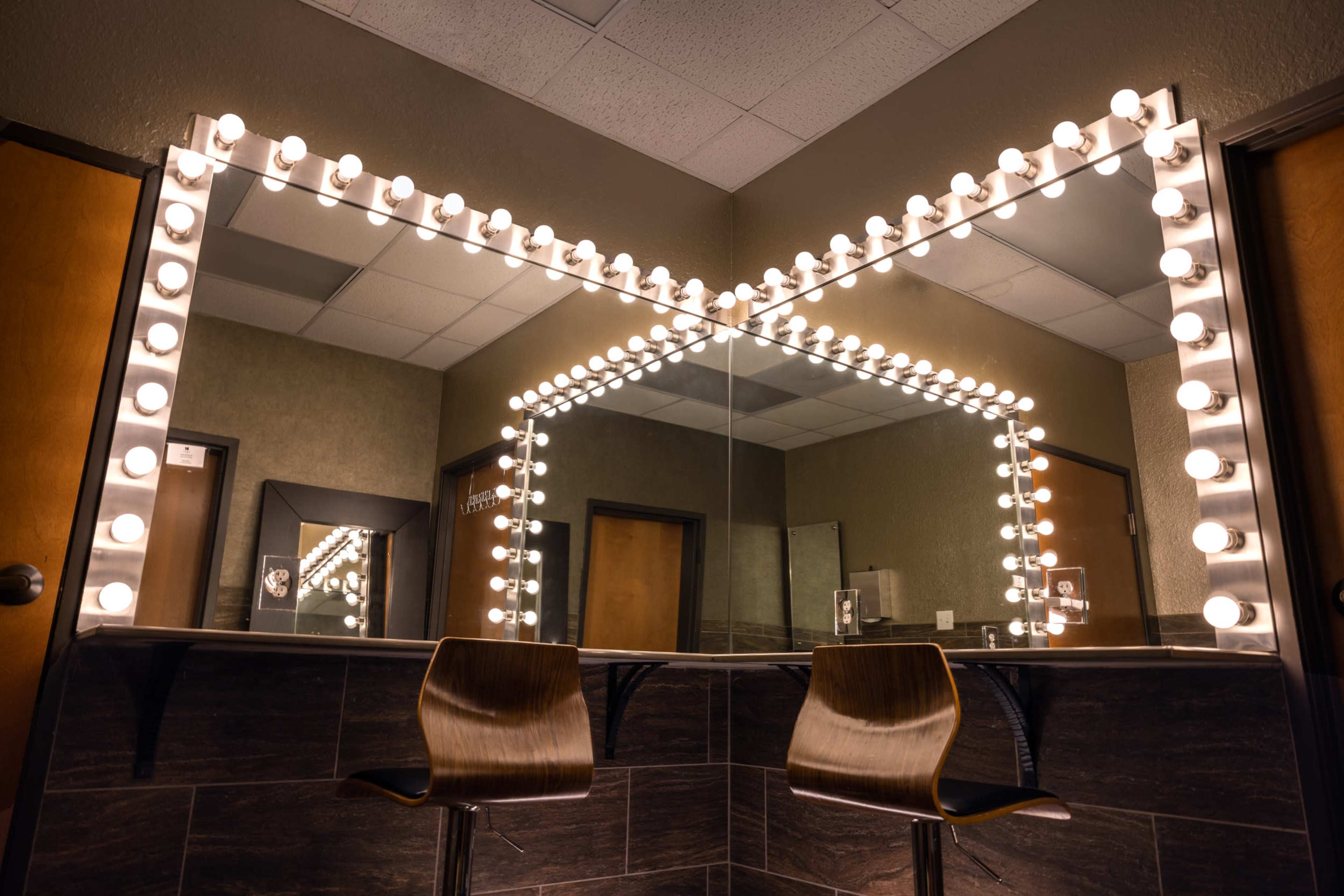 A well-lit vanity area features a large mirror surrounded by light bulbs and two wooden chairs.