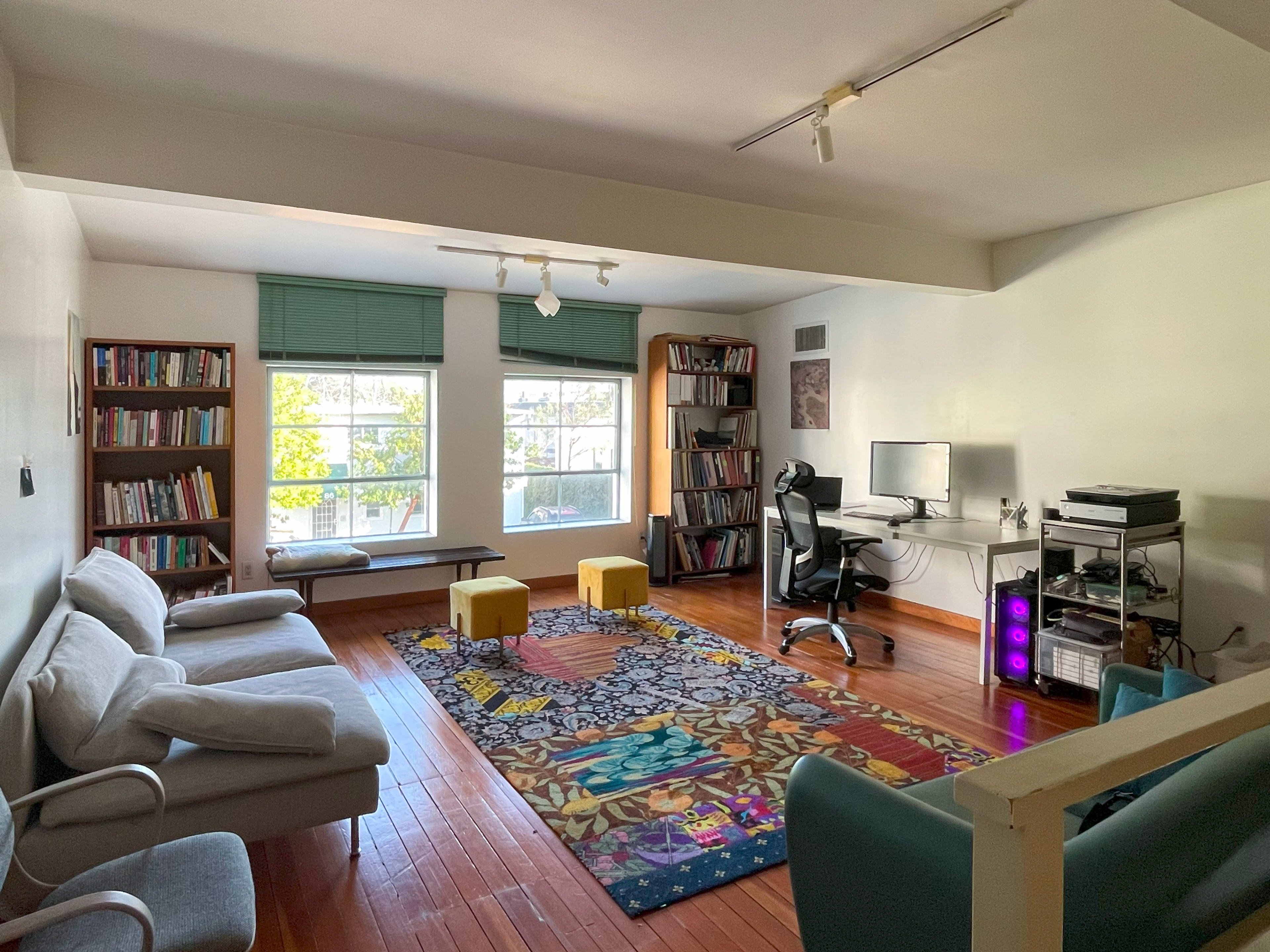 The room features a hardwood floor with a large area rug, a desk with a computer, a bookshelf filled with books, a seating area with two sofas, and two yellow stools.