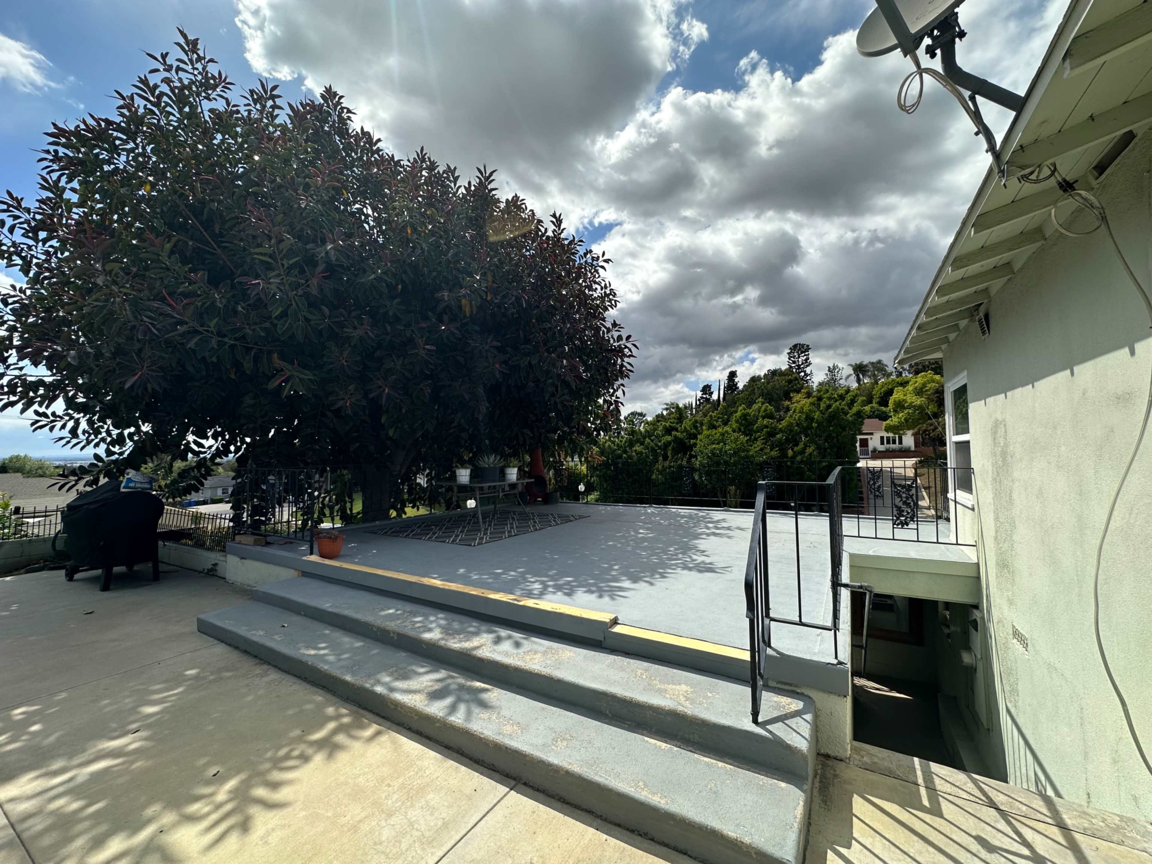 A spacious outdoor area with a large tree, steps leading down, and a house on the right under a partly cloudy sky.