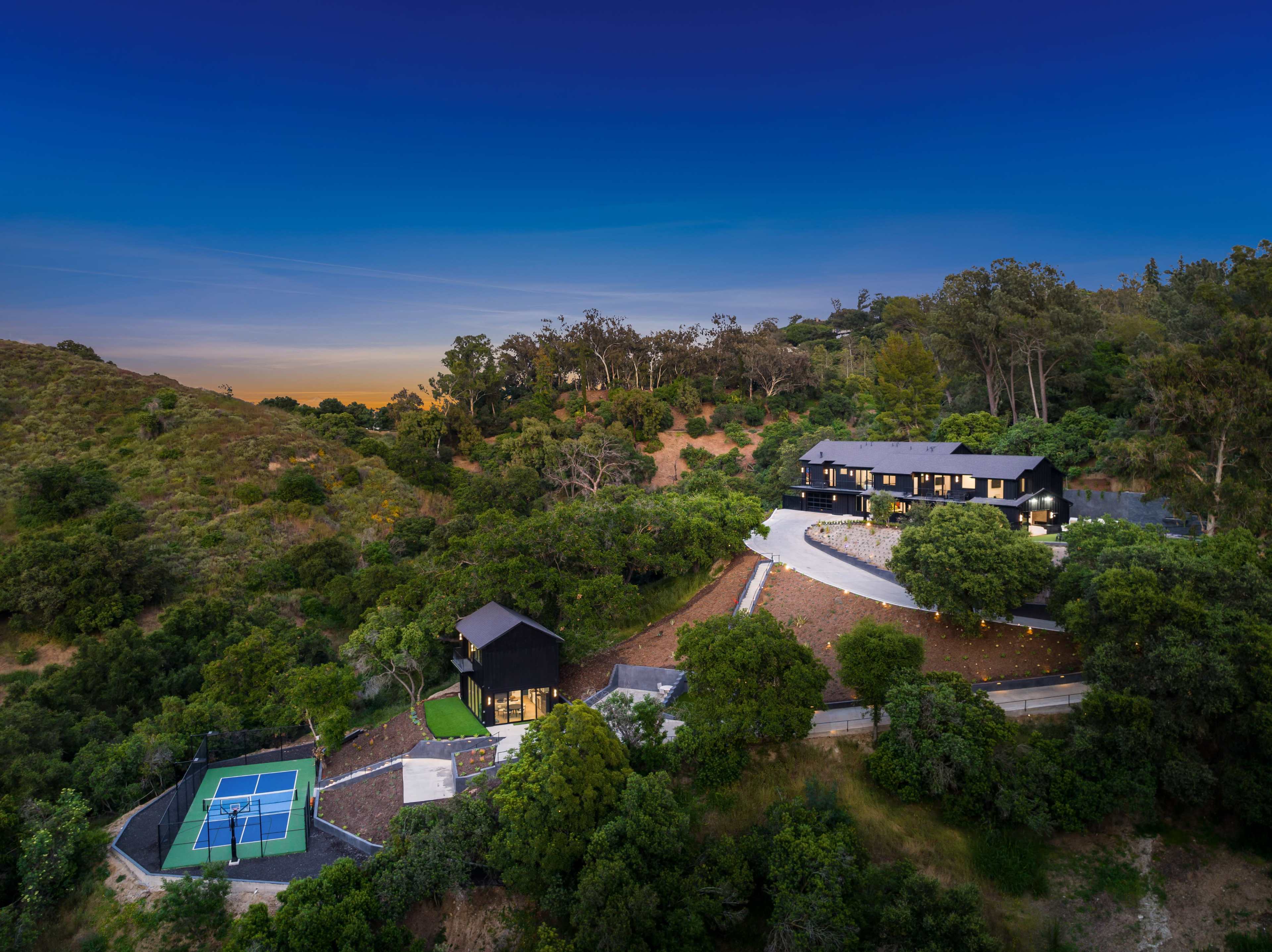 A modern black house is situated on a hillside surrounded by trees, with a tennis court located nearby.