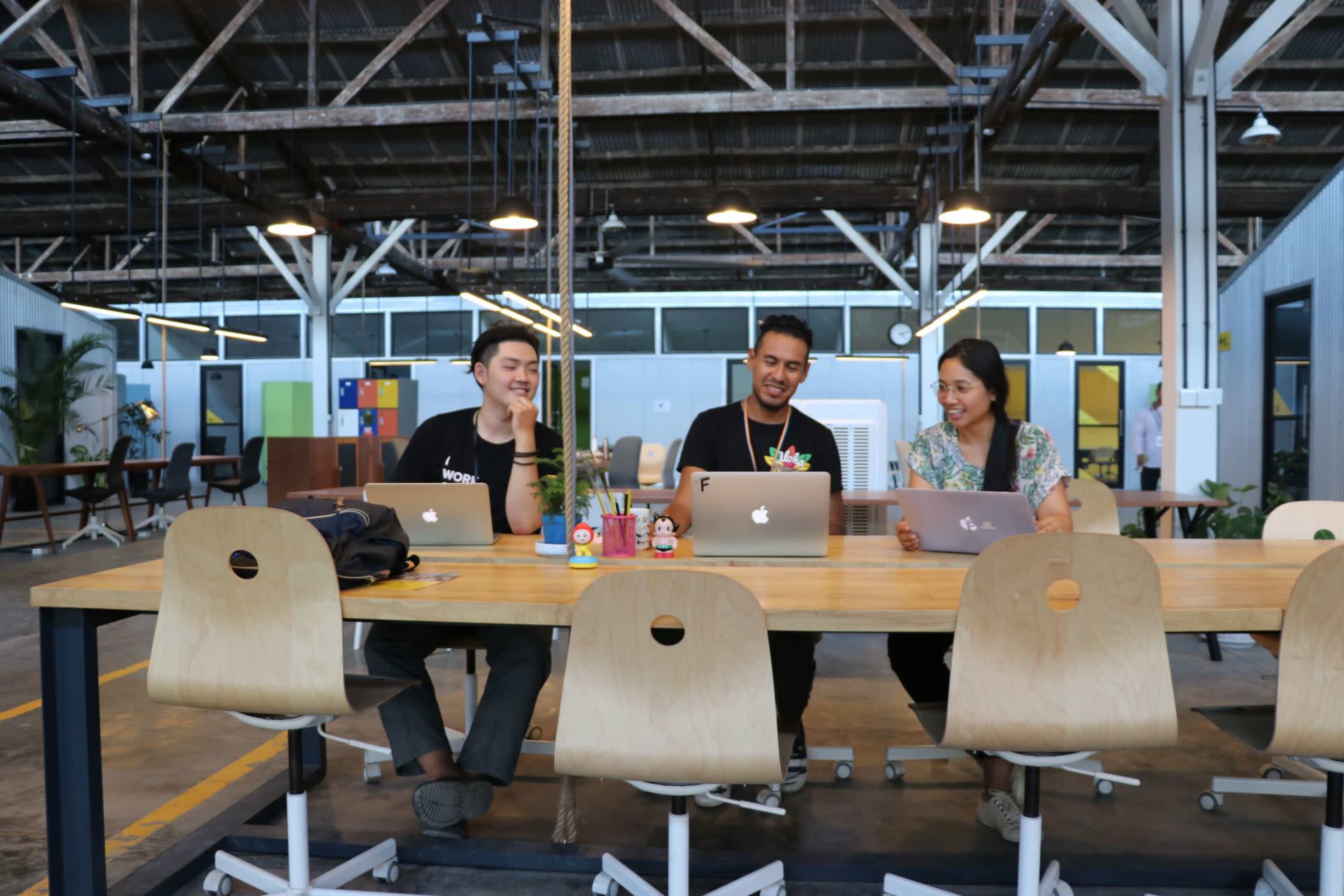 Four people are seated at a wooden table in an open office space, each working on a laptop.