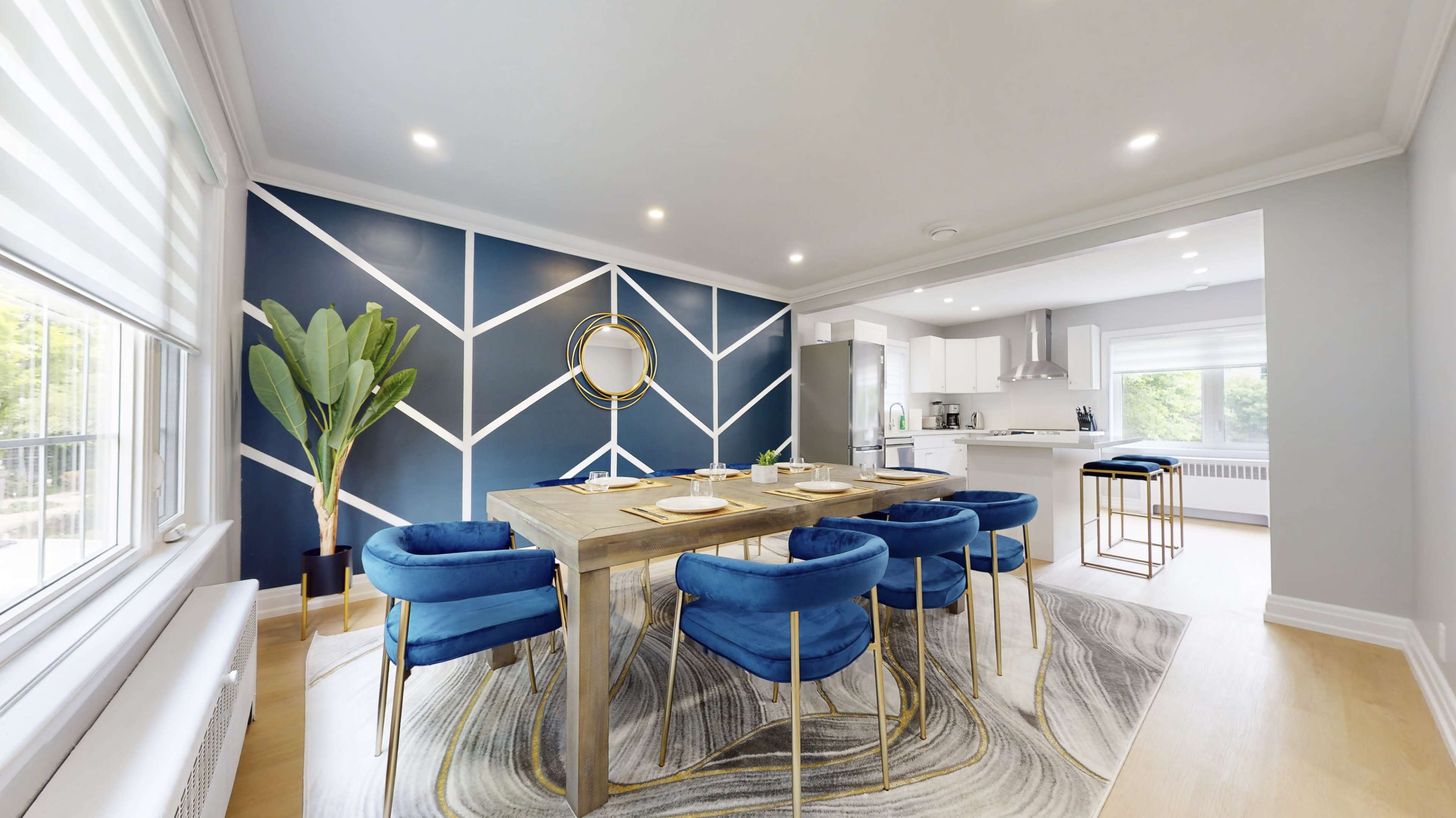 A modern dining area features a large wooden table surrounded by blue velvet chairs, with a geometric accent wall and a kitchen in the background.