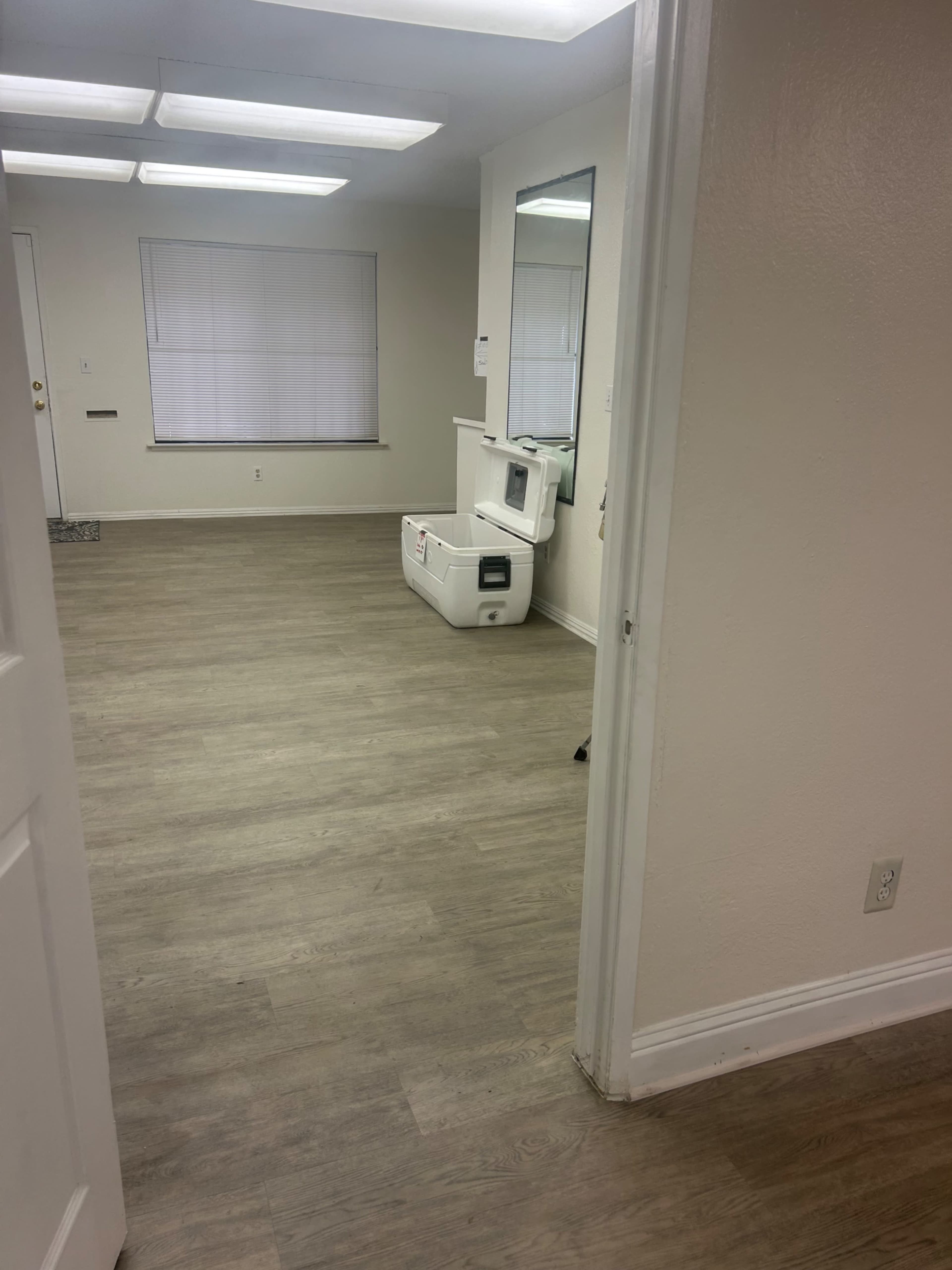 The image shows a spacious, empty room with light-colored walls, a large window, and a cooler positioned on the floor.