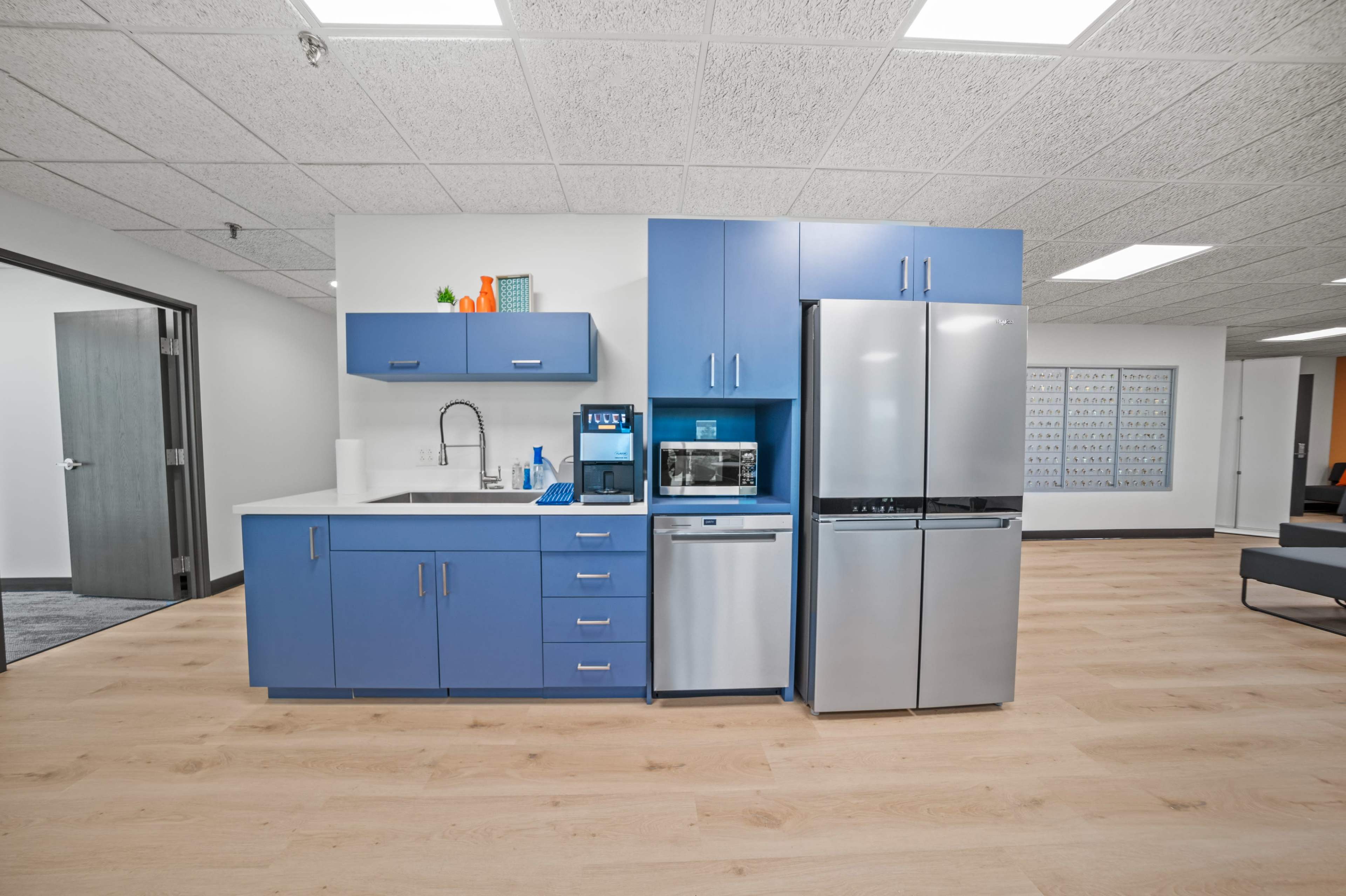 The image shows a modern kitchenette with blue cabinets, a sink, a microwave, a coffee maker, and stainless steel appliances, all situated in a well-lit office space.