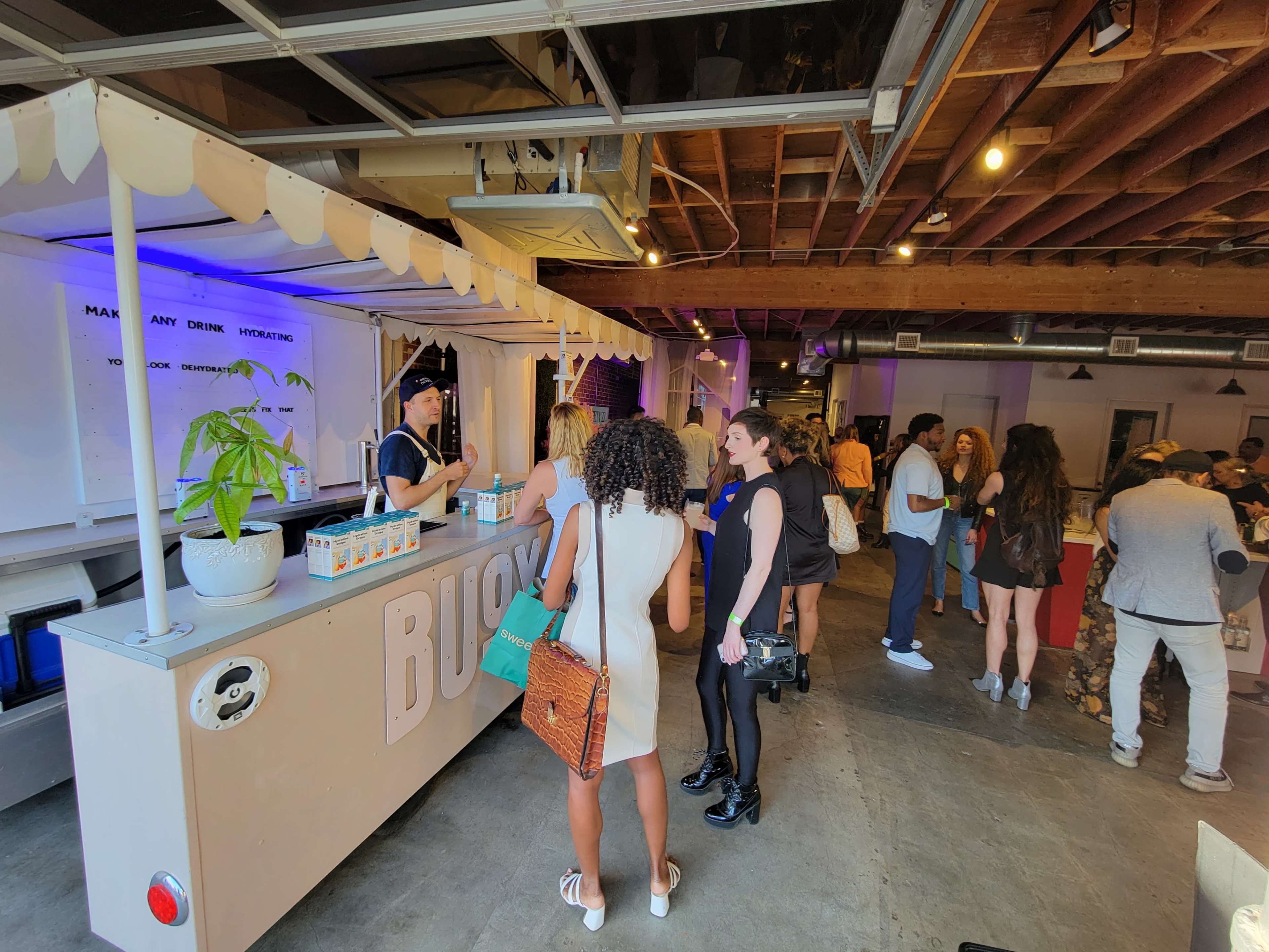 A group of people gathers around a mobile bar setup inside a spacious indoor venue, engaging in conversation and enjoying drinks.