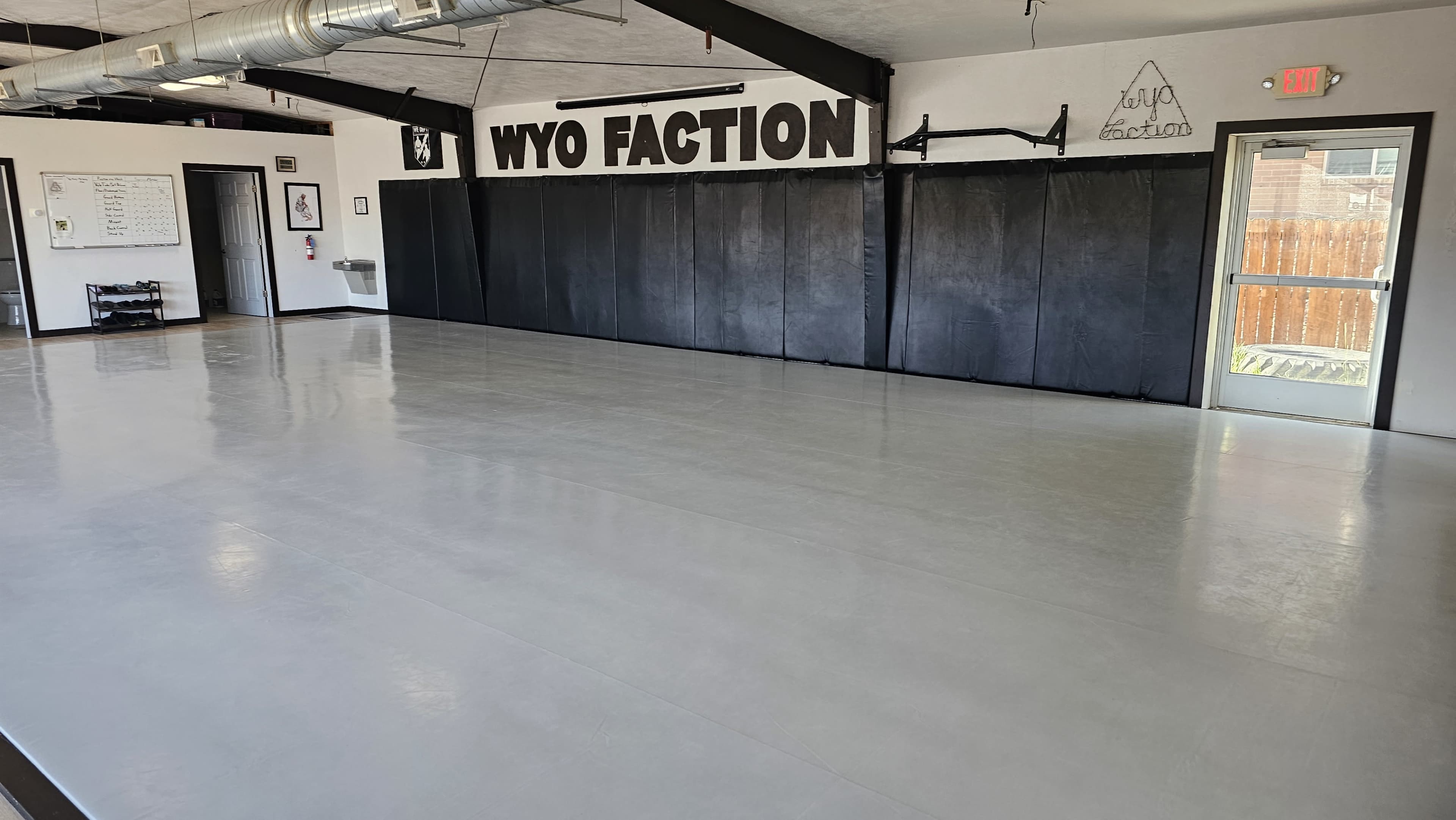 The image shows the interior of a martial arts training room with matte gray flooring, black padded walls, and minimal furniture.