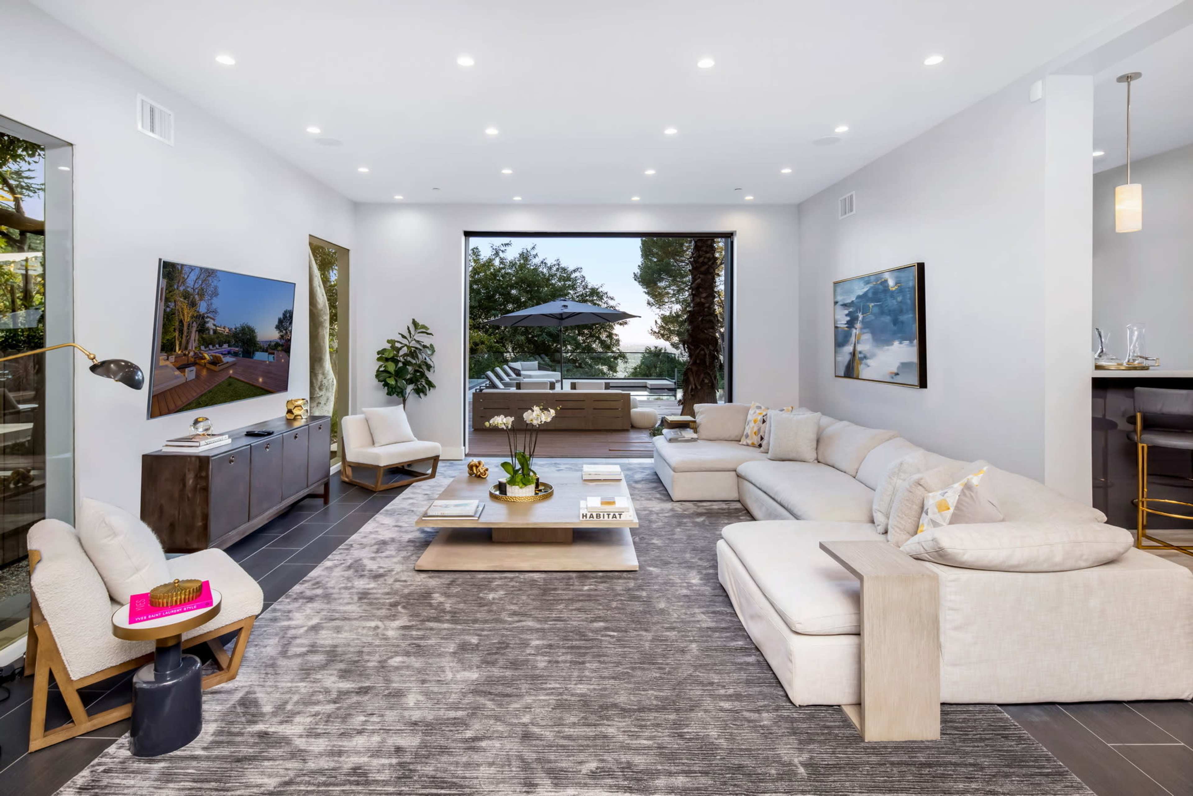The image shows a contemporary living room with large windows, a light-colored sectional sofa, and a flat-screen TV mounted on the wall.