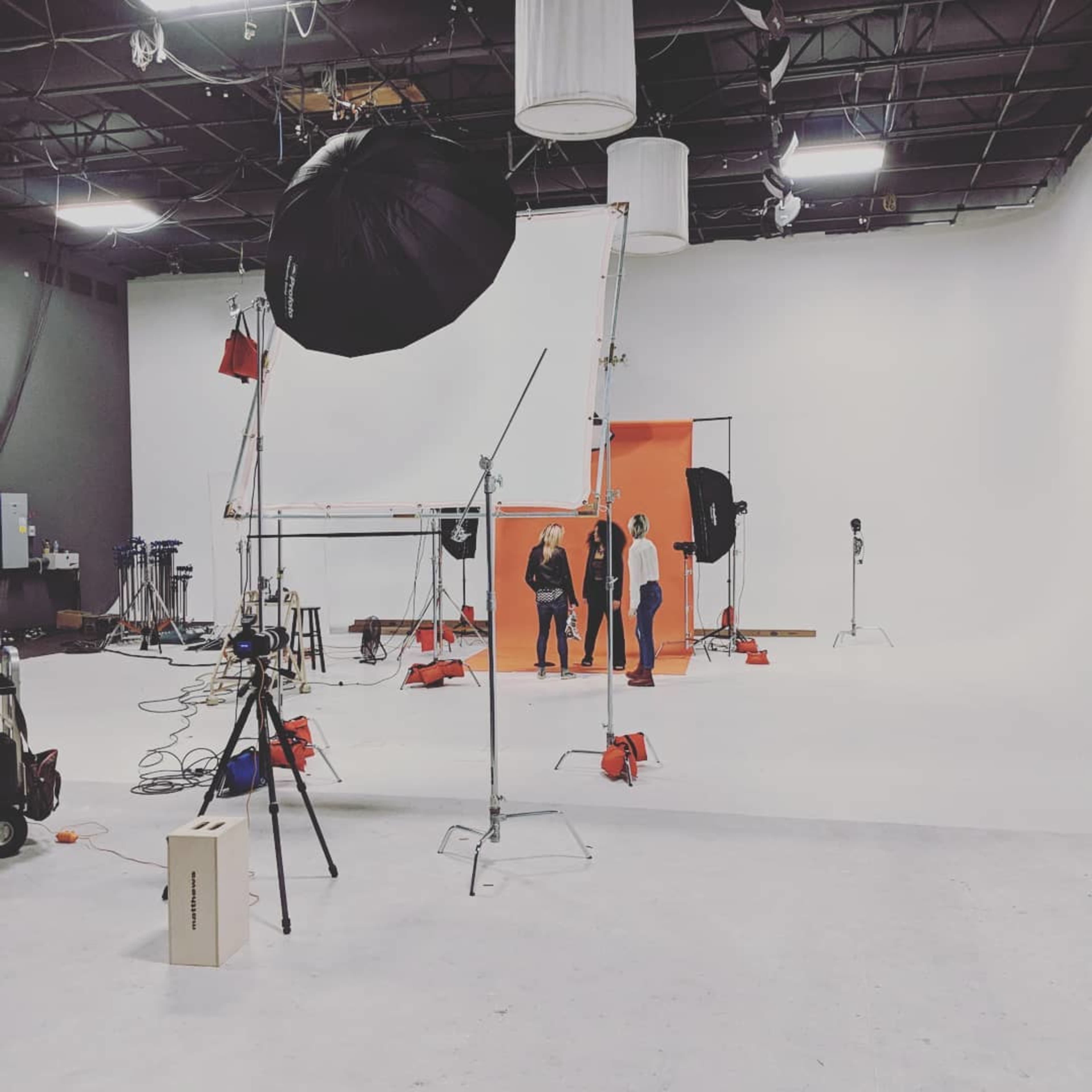 A photography studio features an orange backdrop with two individuals positioned in front of it, surrounded by various lighting equipment and support stands.