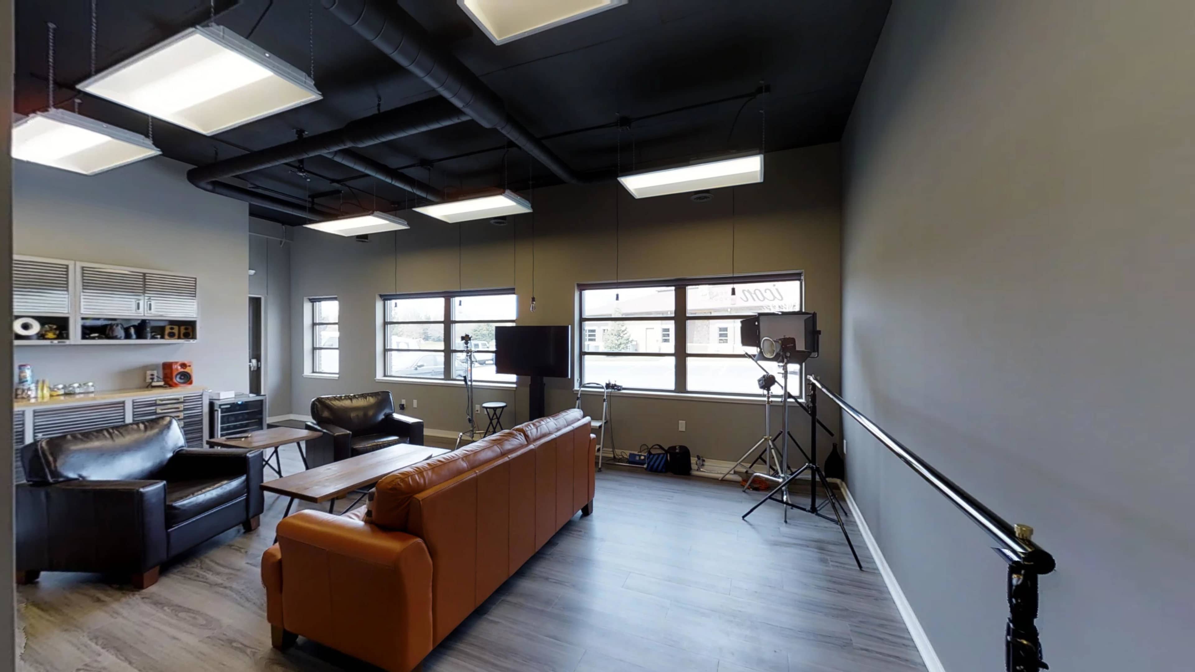 A modern lounge area with a brown leather sofa, two black chairs, a wooden coffee table, and equipment for media production, all set against a gray wall and large windows.