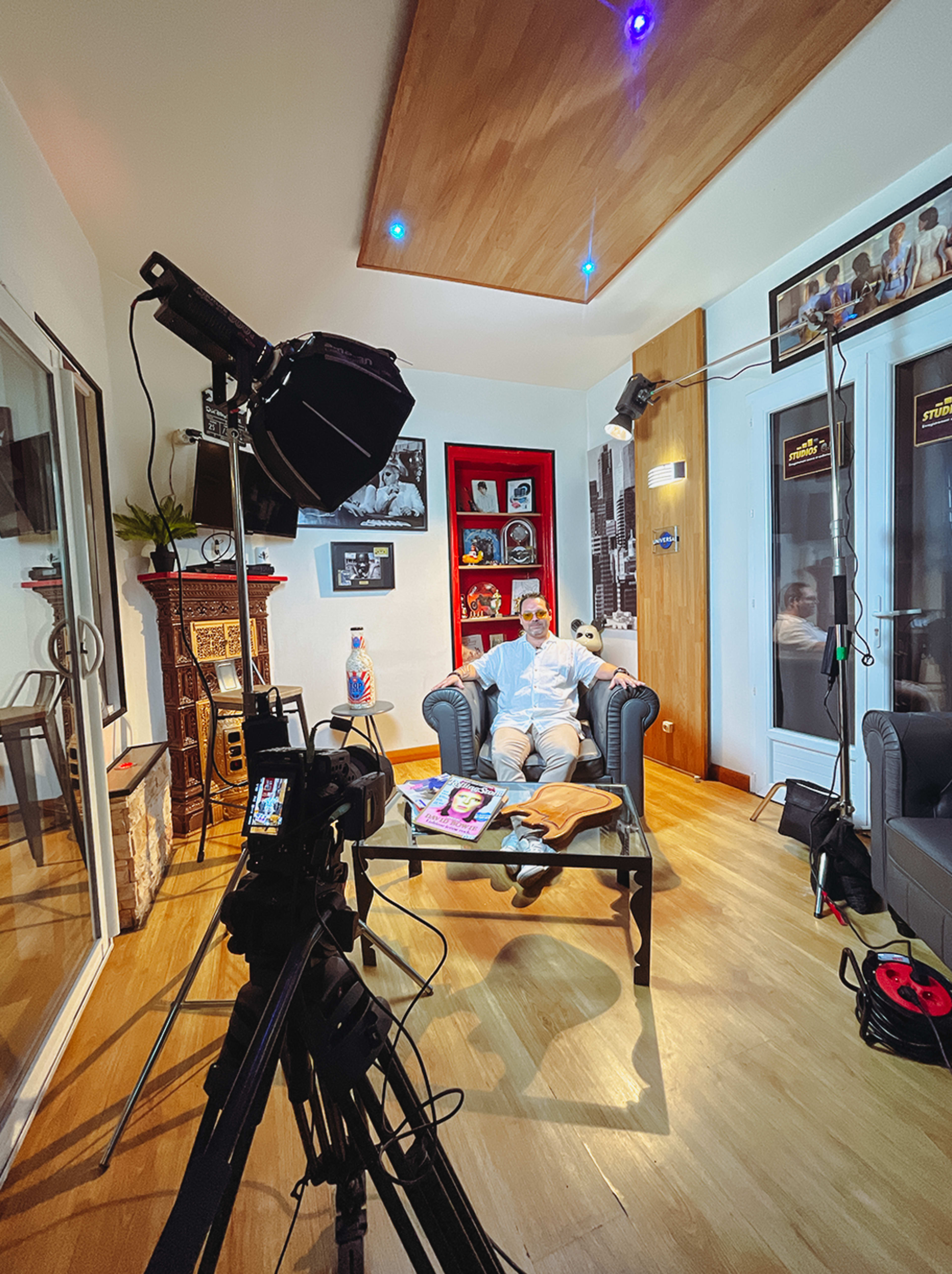 A man sits on a couch in a well-decorated room, surrounded by various objects and a camera setup pointing towards him.