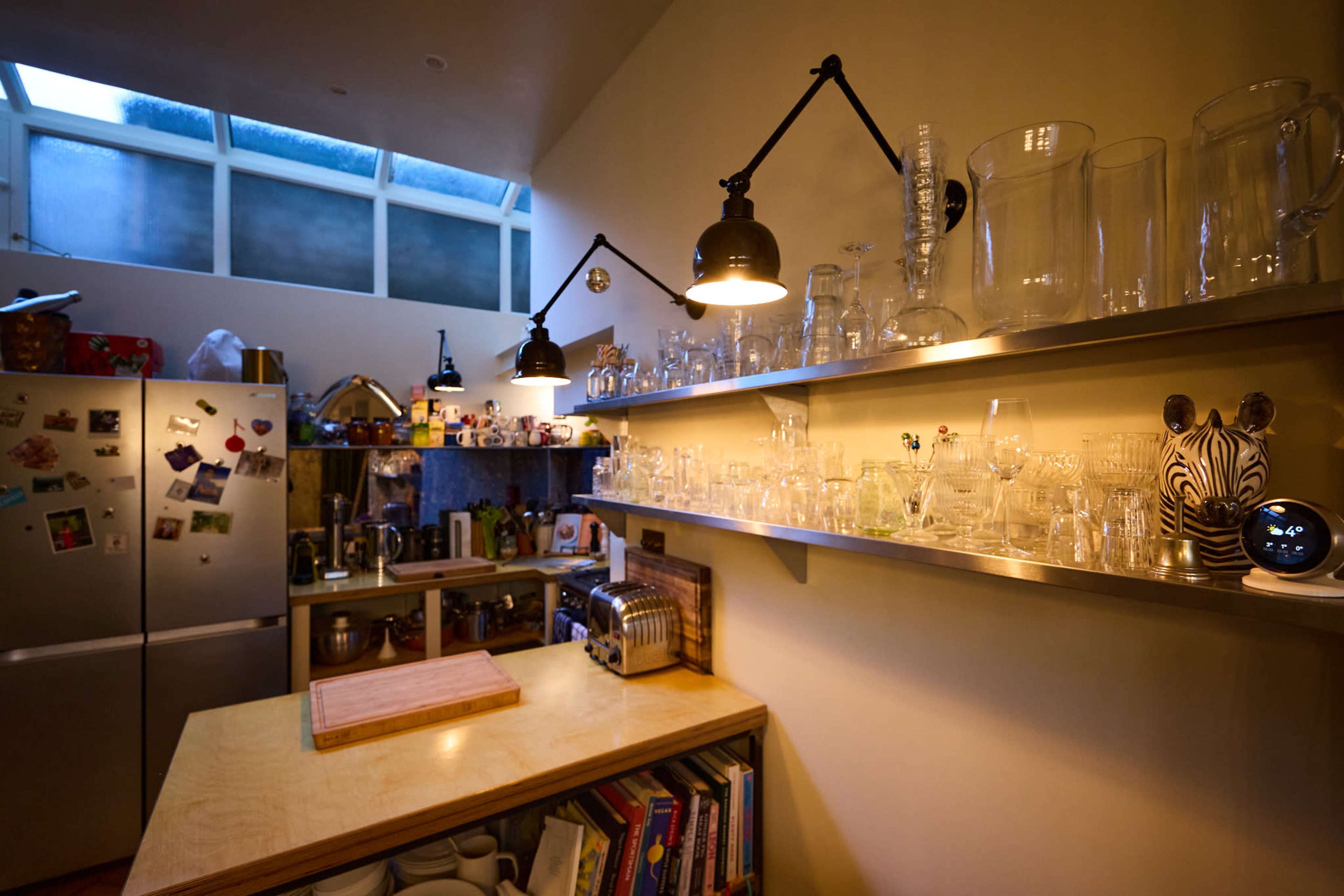 The kitchen features a wooden countertop with various kitchen items and a shelf displaying an assortment of glassware under warm lighting.