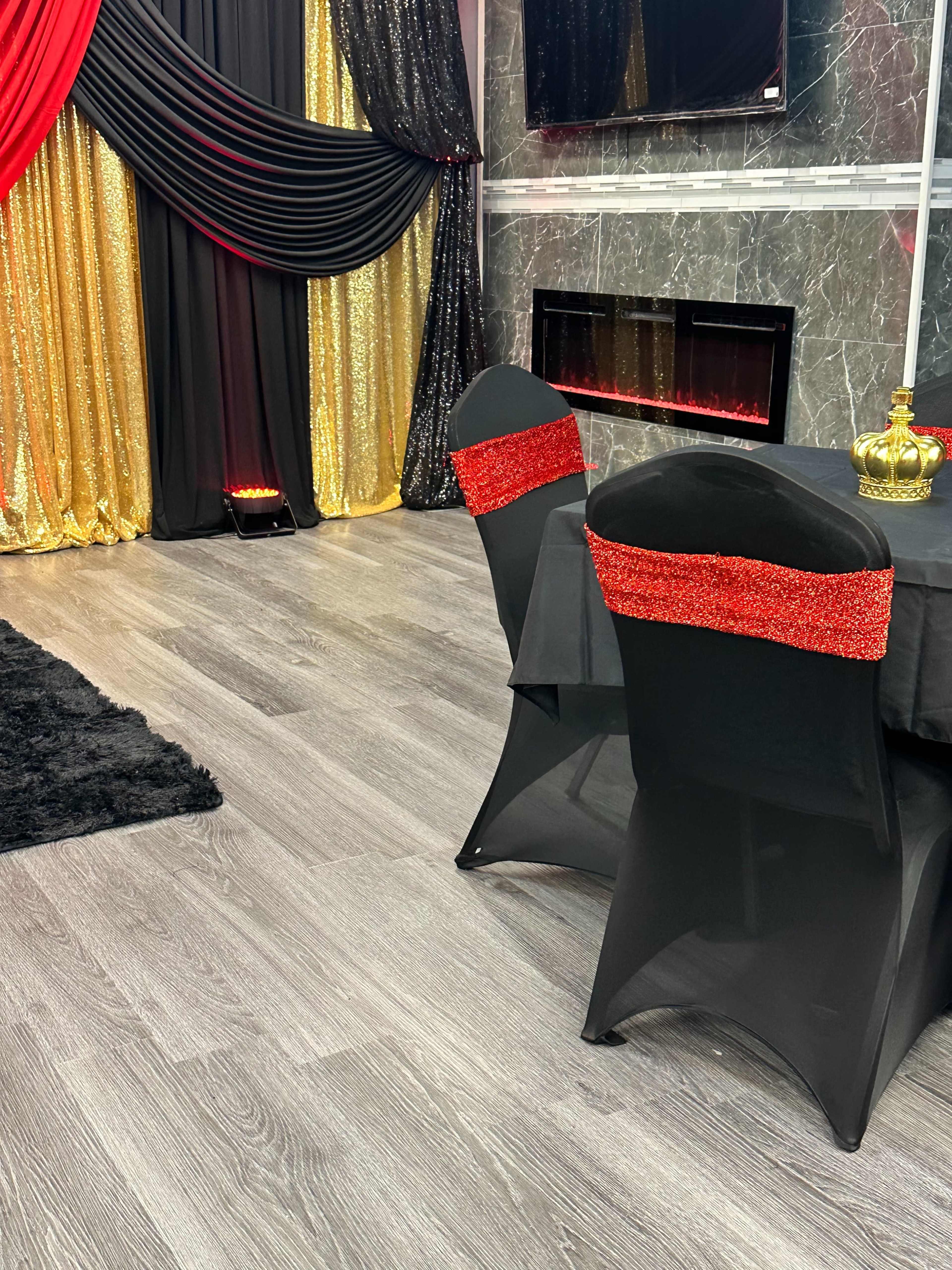 A room with a black table covered with a sparkly red and black table runner, flanked by two black chairs adorned with red sequined sashes, against a backdrop of gold and black curtains.