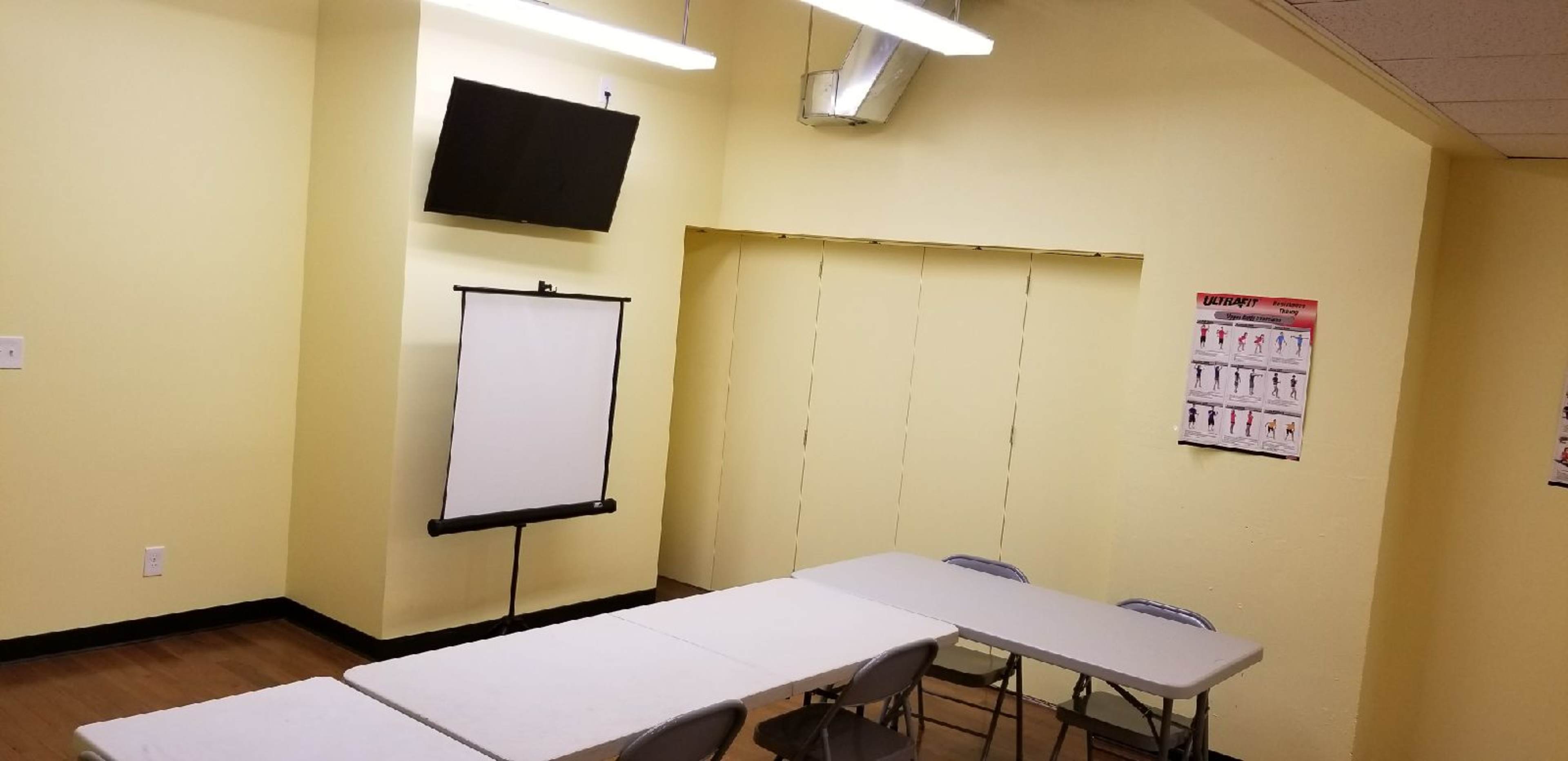A small, empty meeting room with a yellow wall, a mounted TV, a presentation board, and a row of folding tables and chairs.